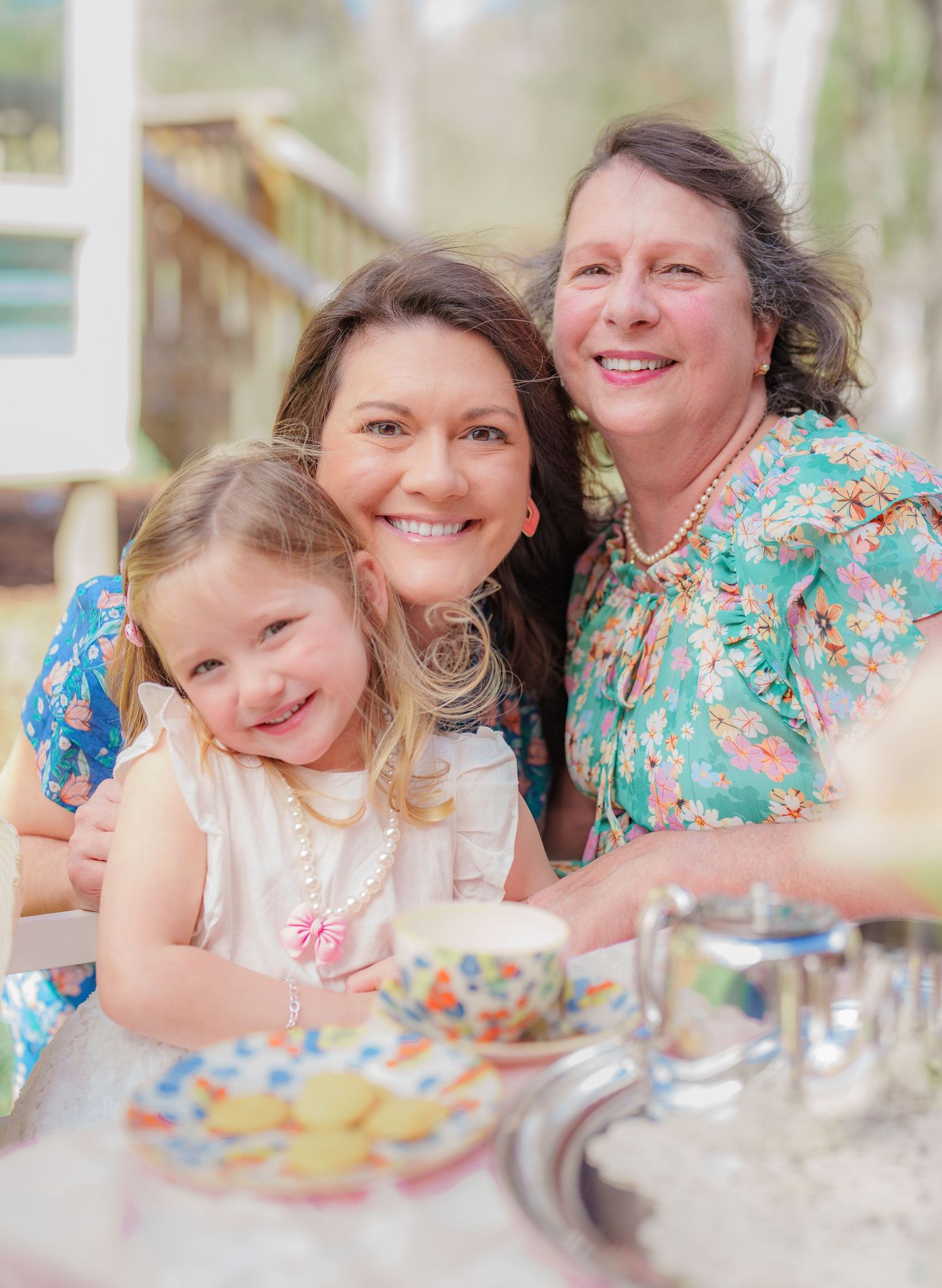 Three generations of women smile together at a colorful tea party table outdoors.