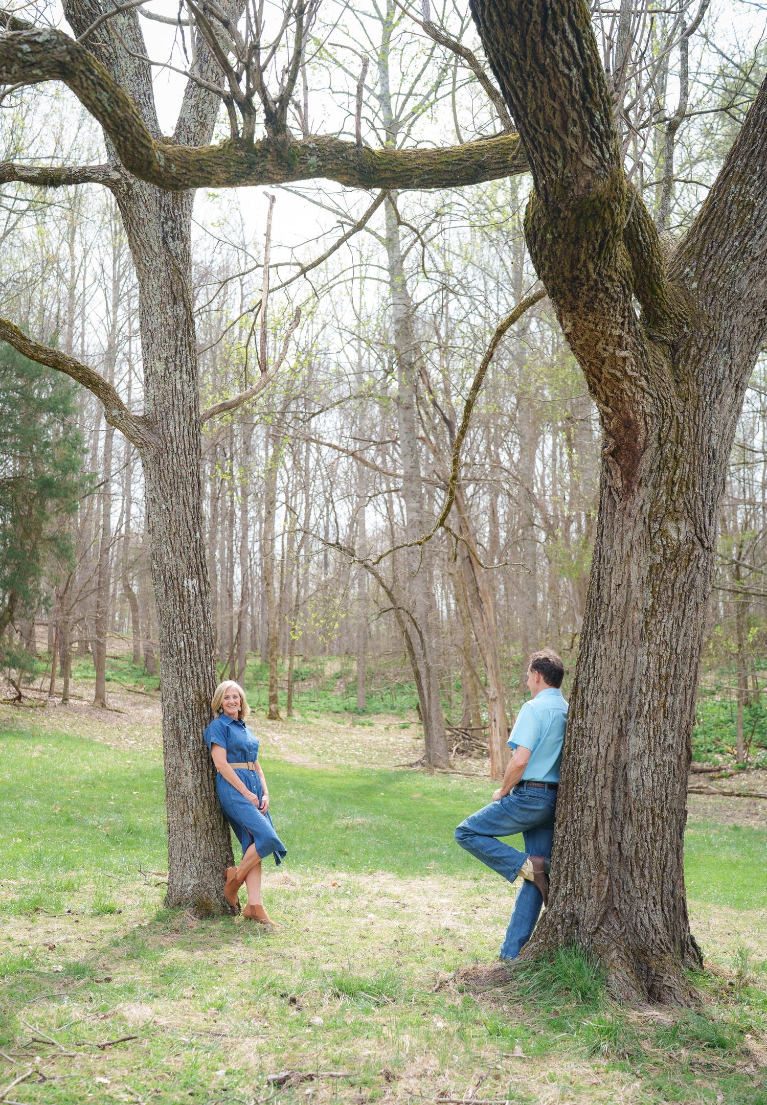 Couple leaning against separate trees in a wooded area, smiling at each other playfully.
