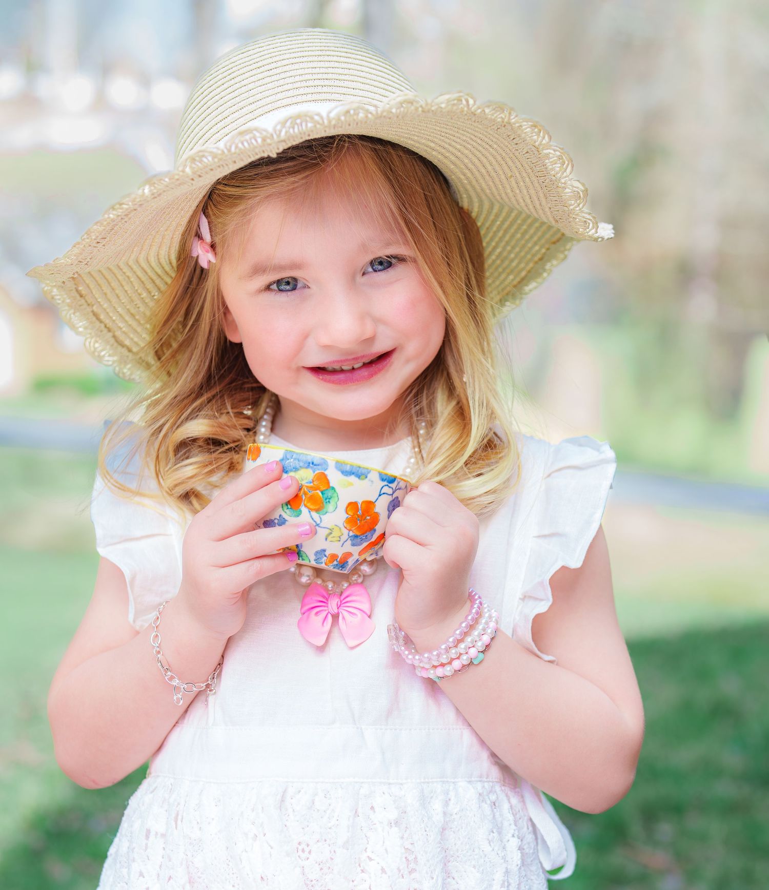 Smiling little girl in white dress and straw sun hat holding a floral teacup outdoors.