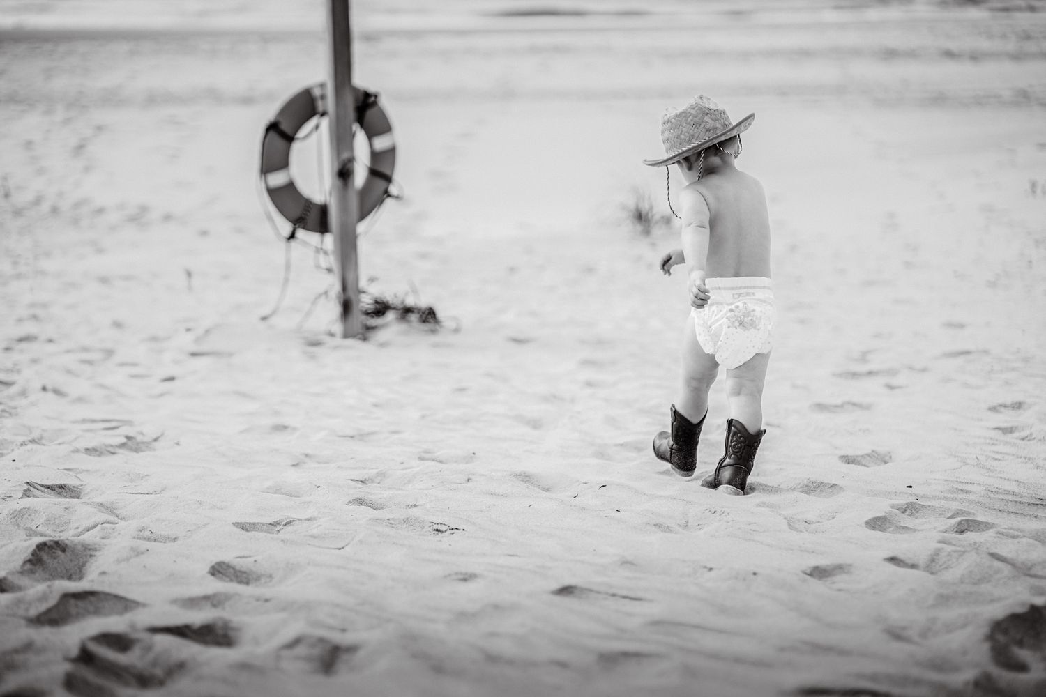 Toddler in cowboy hat and boots walks on sandy beach near life preserver ring pole.