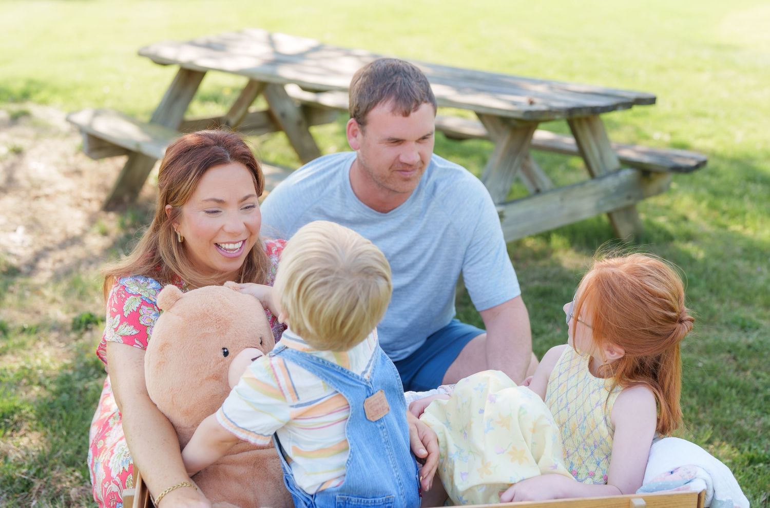 Happy family sitting on grass at picnic area with young children and teddy bear on sunny day.
