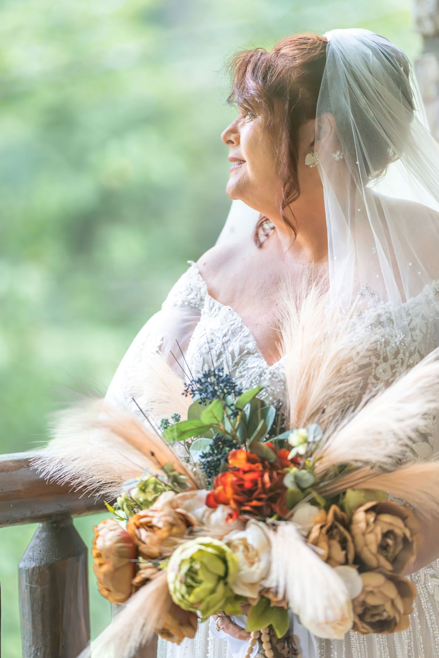 Bride in white gown and veil holds a rustic bouquet with roses and pampas grass on a wooden balcony.