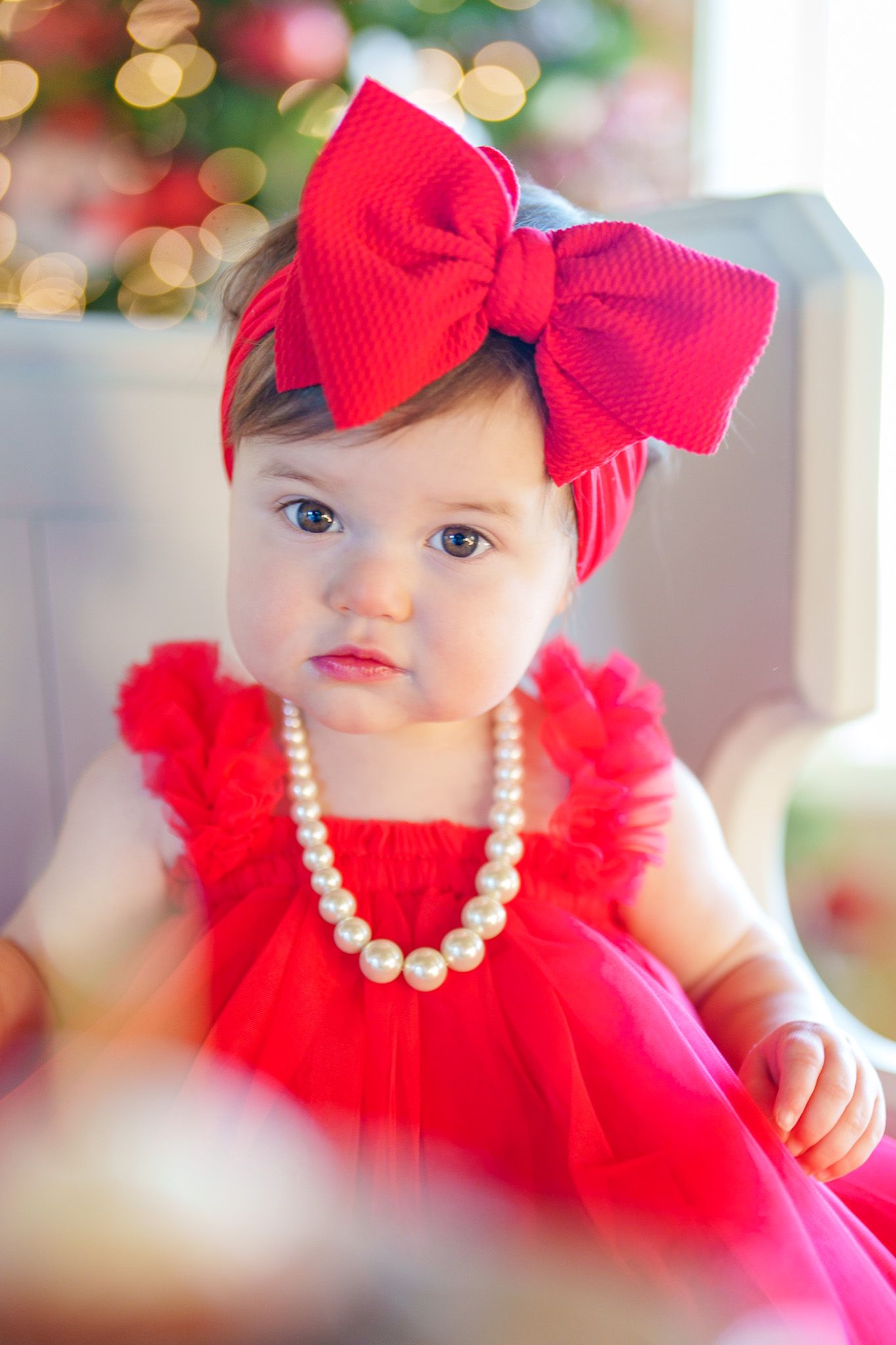 Adorable baby girl in red tulle dress with large bow headband and pearl necklace near Christmas tree.