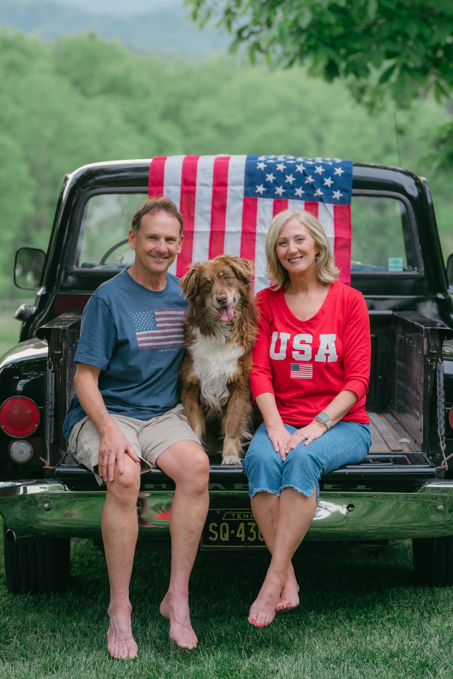 Smiling couple in patriotic attire sitting with their dog in a vintage truck bed.