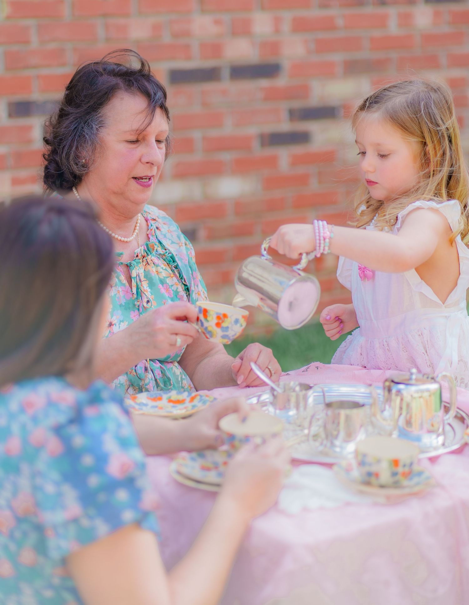 Young girl pours tea at outdoor table with two women during a charming garden tea party.