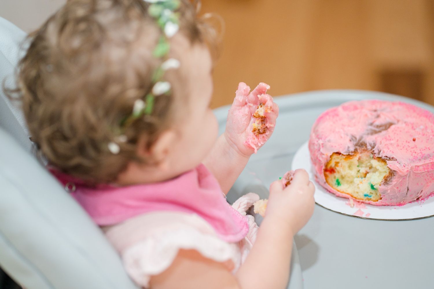 Baby girl with floral crown digging into a pink smash cake during her first birthday celebration.