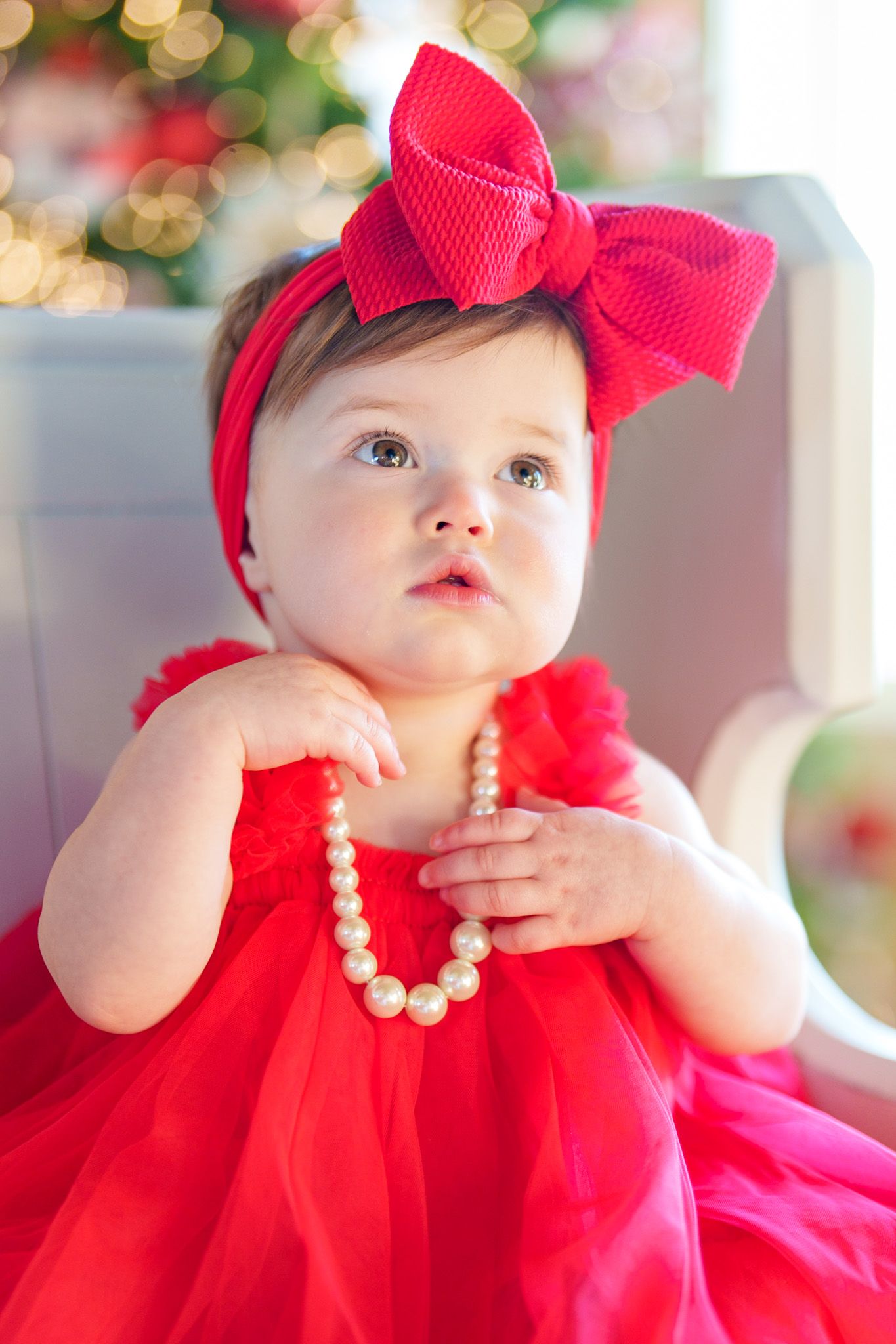 Adorable baby girl in red dress with large bow headband holding pearl necklace near Christmas tree.