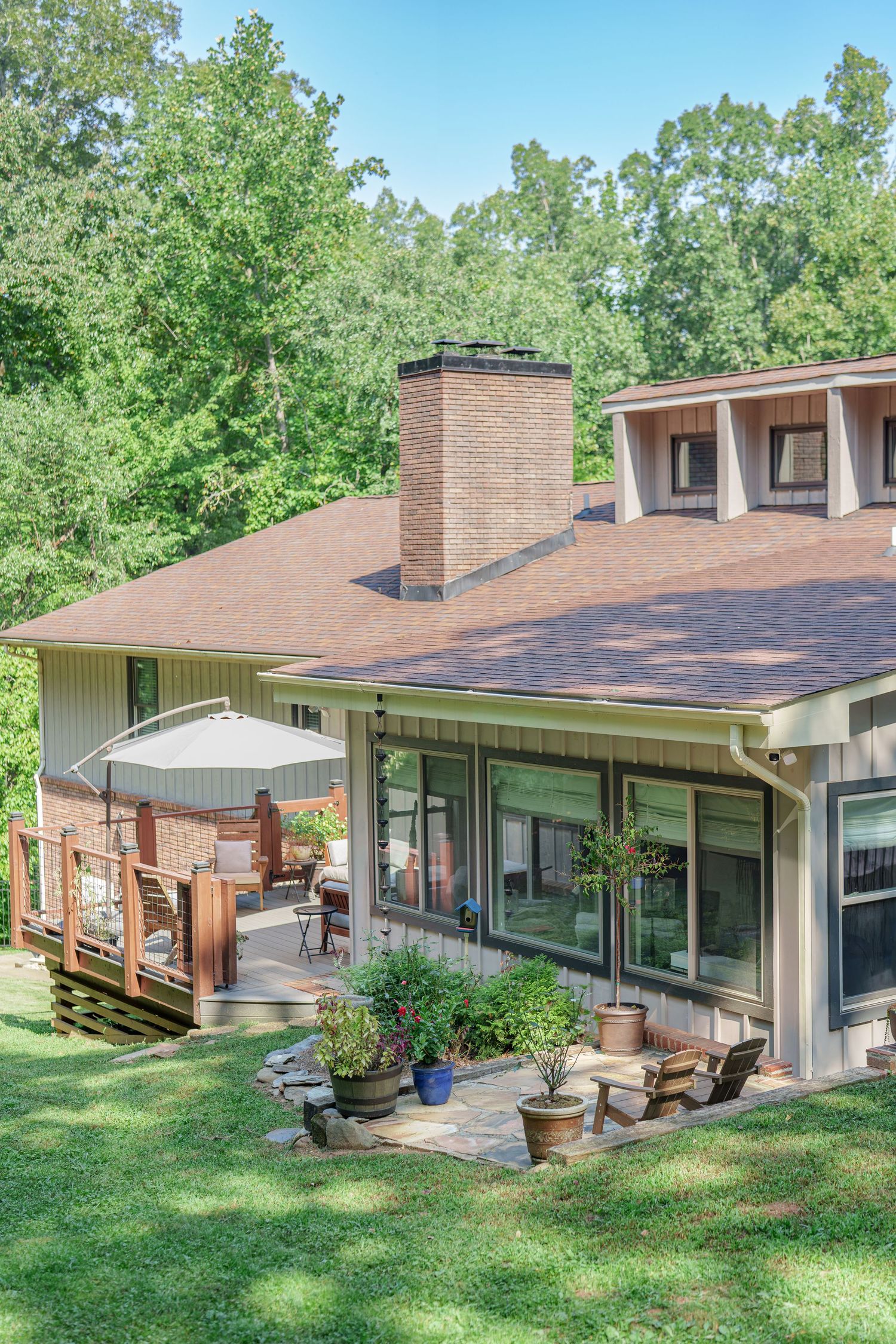 Aerial view of a charming home with brick chimney, wood deck, patio seating, and lush green trees in the background.