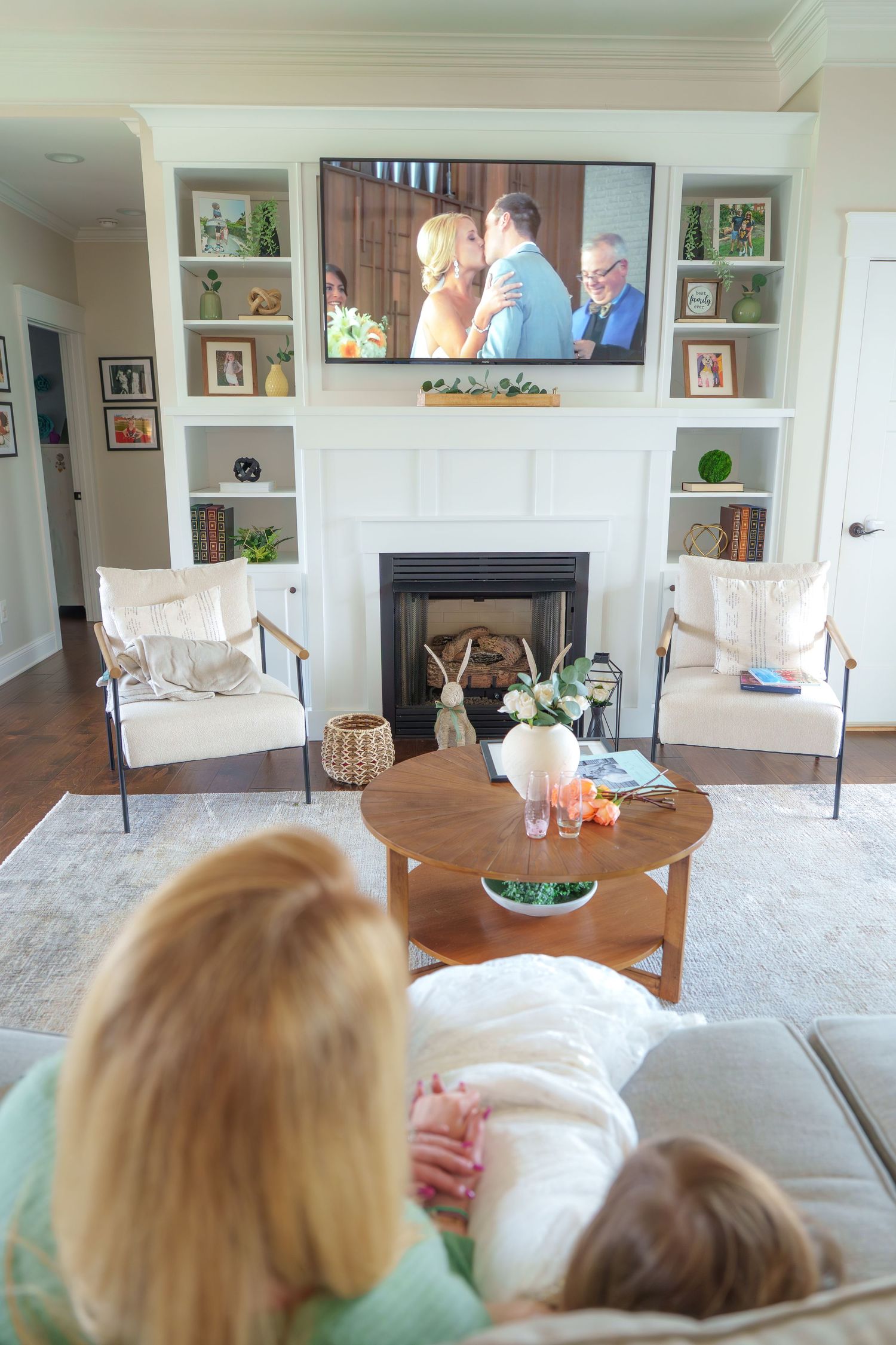 Two people relaxing on a couch watching TV above a white fireplace with built-in shelves in a cozy living room.