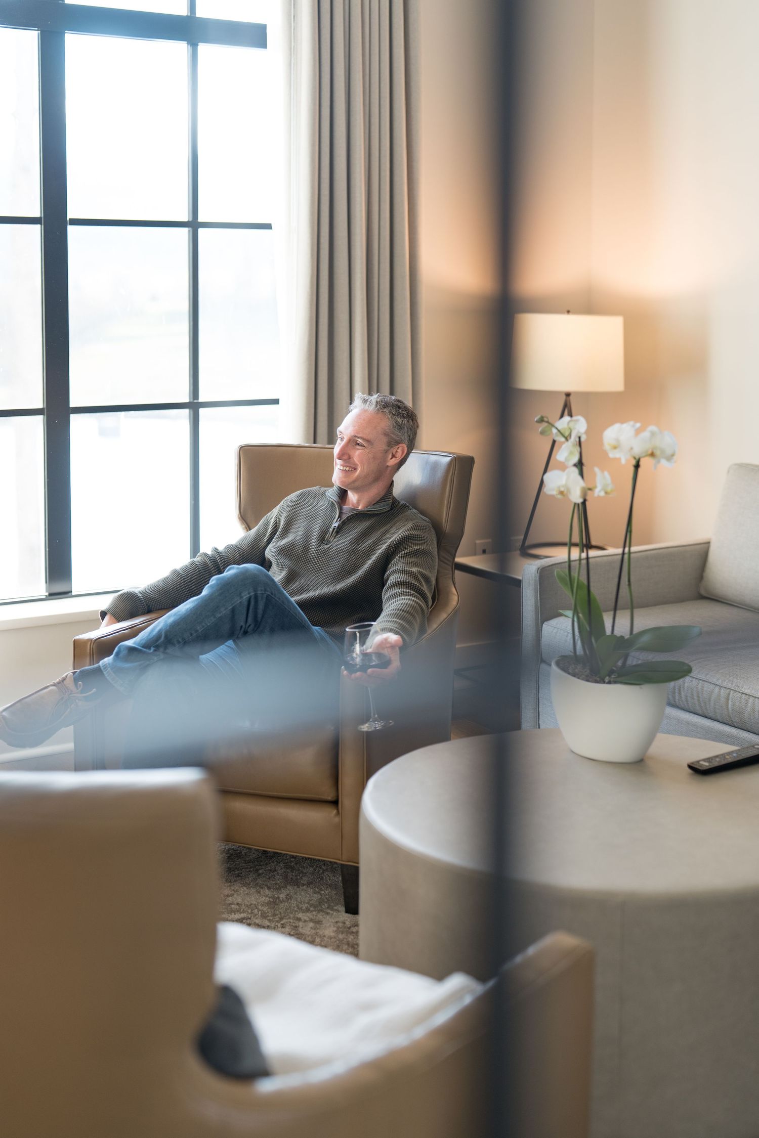 A relaxed middle-aged man sitting in a leather chair by a window in a modern hotel suite, smiling contentedly.