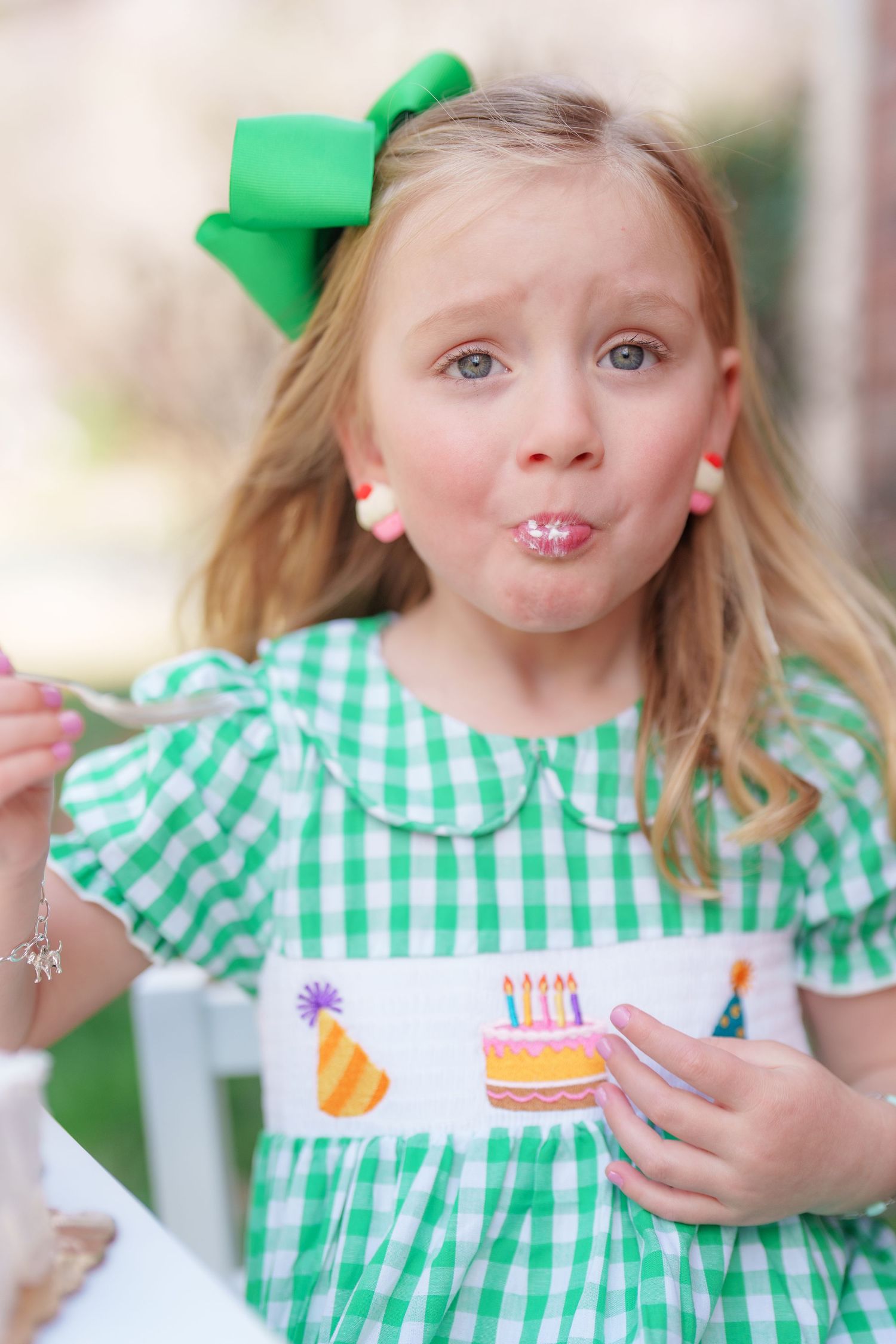 Young girl in green gingham smocked birthday dress with bow, eating cake at a birthday party.