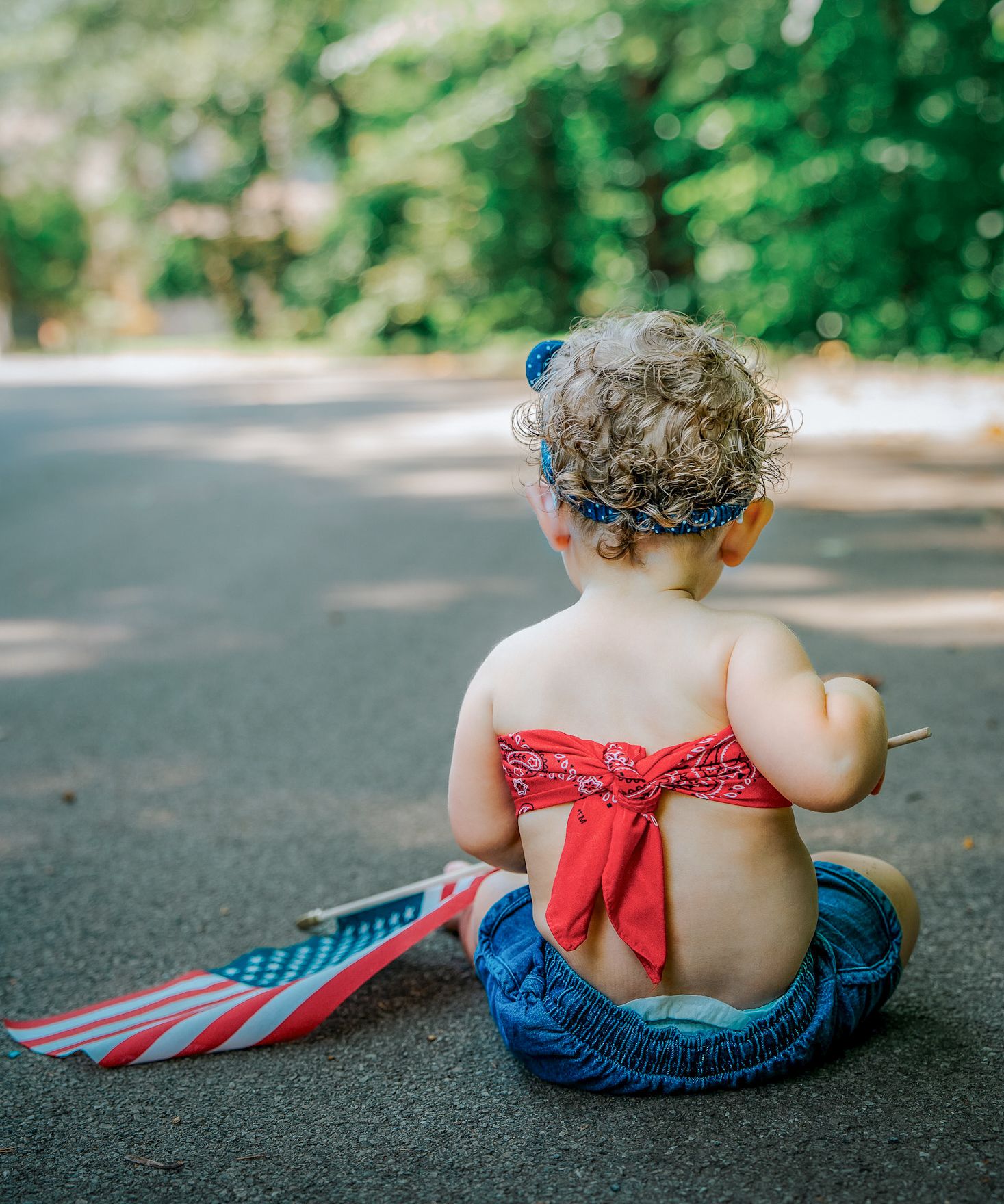 A curly-haired toddler sits on a path holding an American flag, wearing a patriotic red bandana outfit.