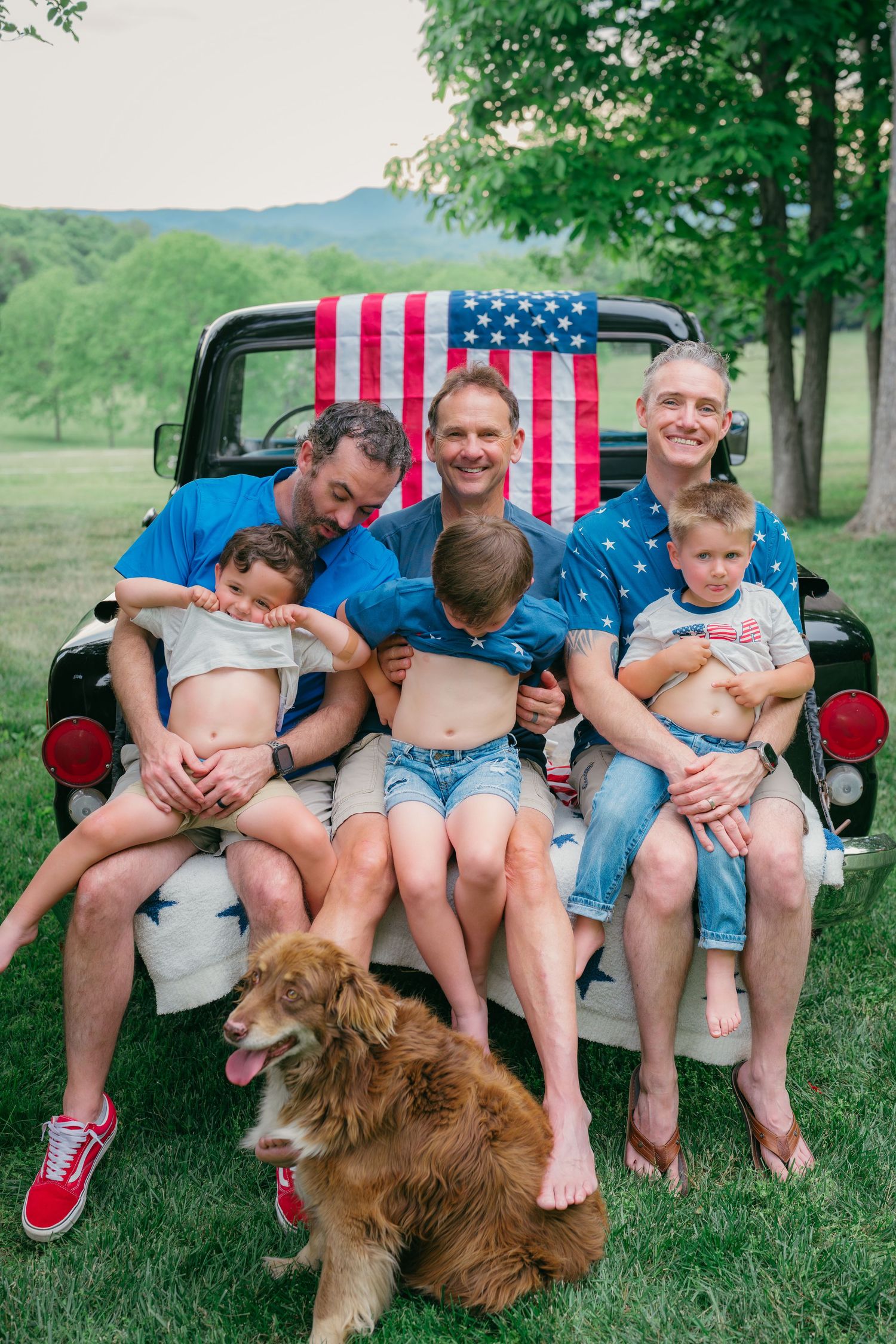 Three men with young children sitting in vintage truck bed with American flag backdrop.