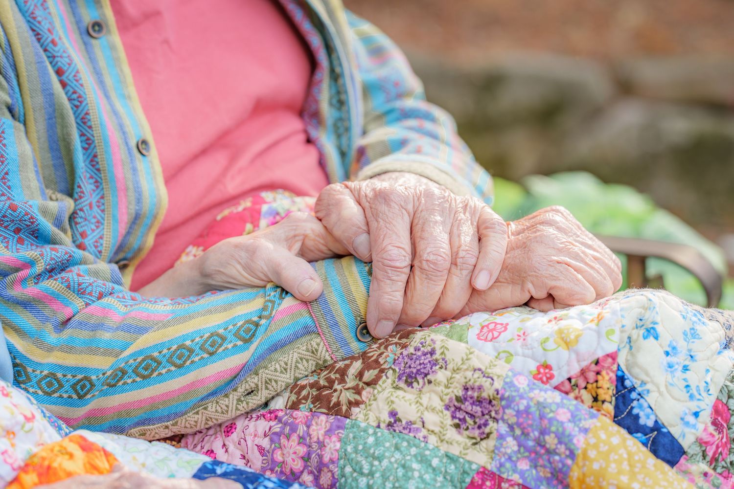 Close-up of elderly hands resting on a colorful patchwork quilt outdoors.