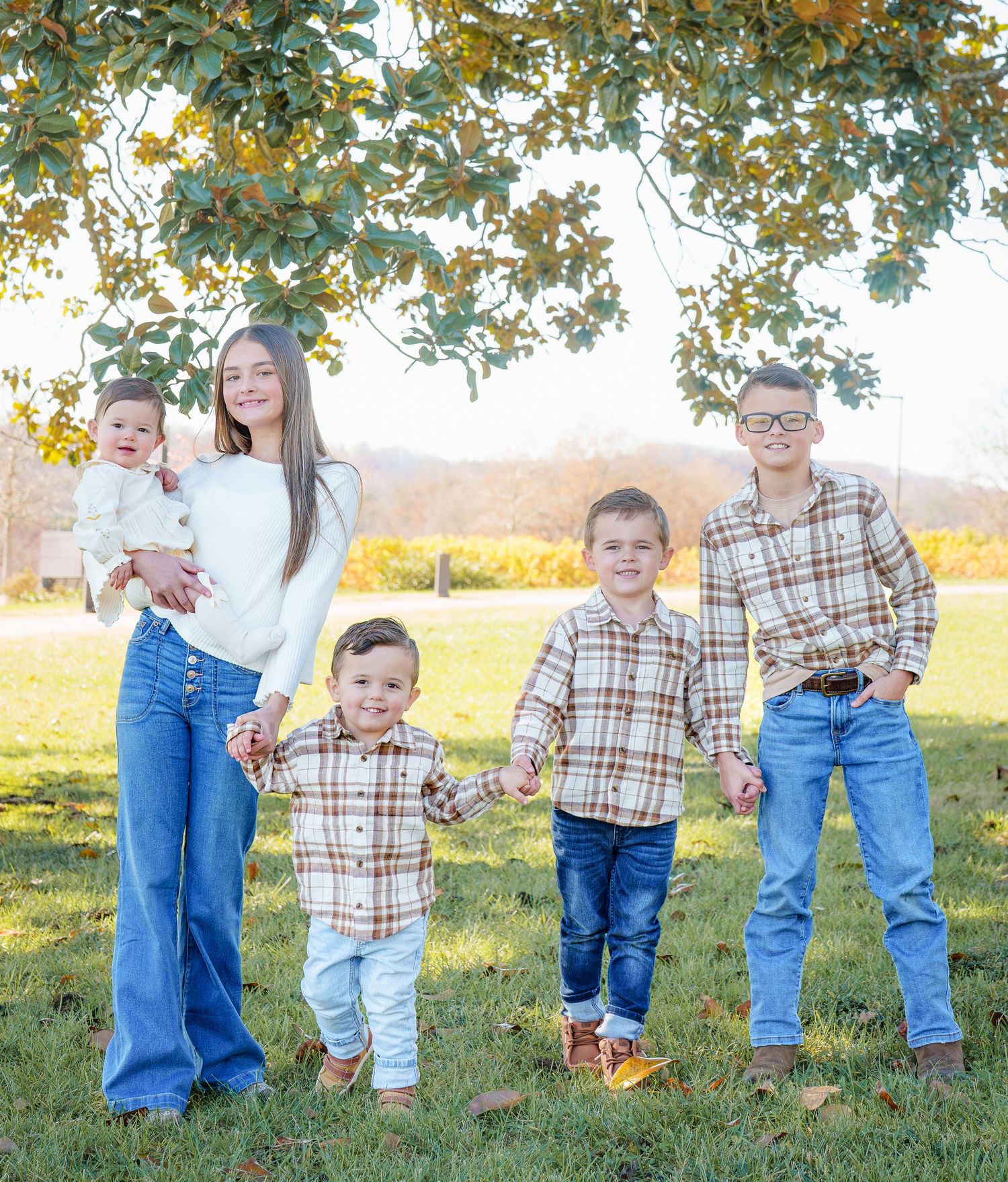 Four siblings in matching plaid shirts and jeans pose together outdoors under a tree with golden autumn foliage.