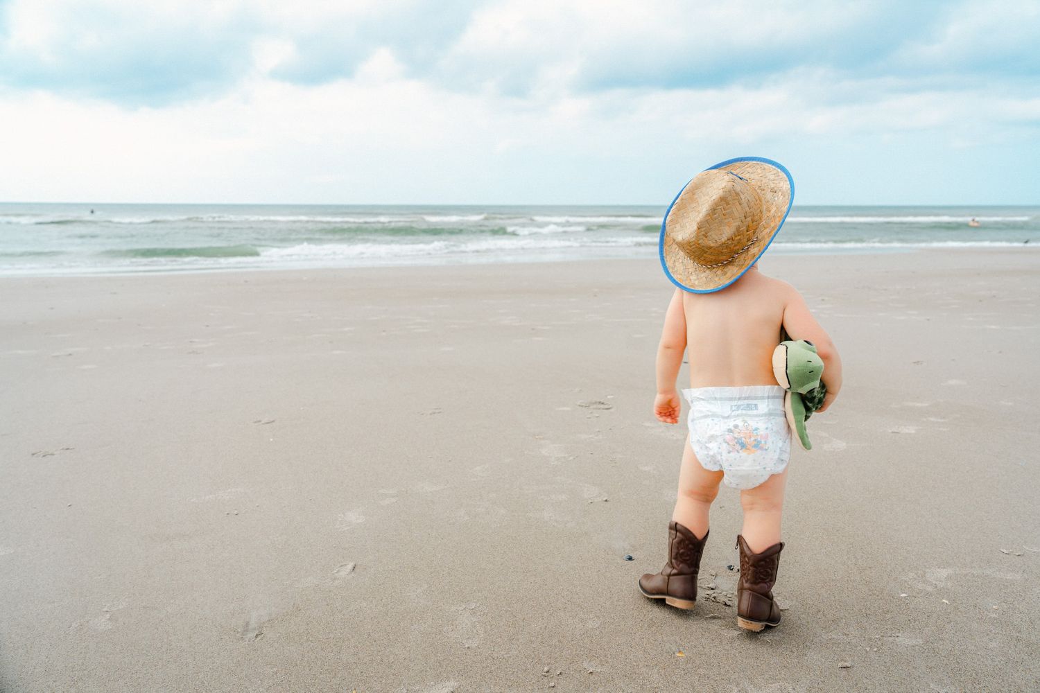Baby boy in cowboy hat, diaper, and boots standing on a sandy beach facing the ocean waves.