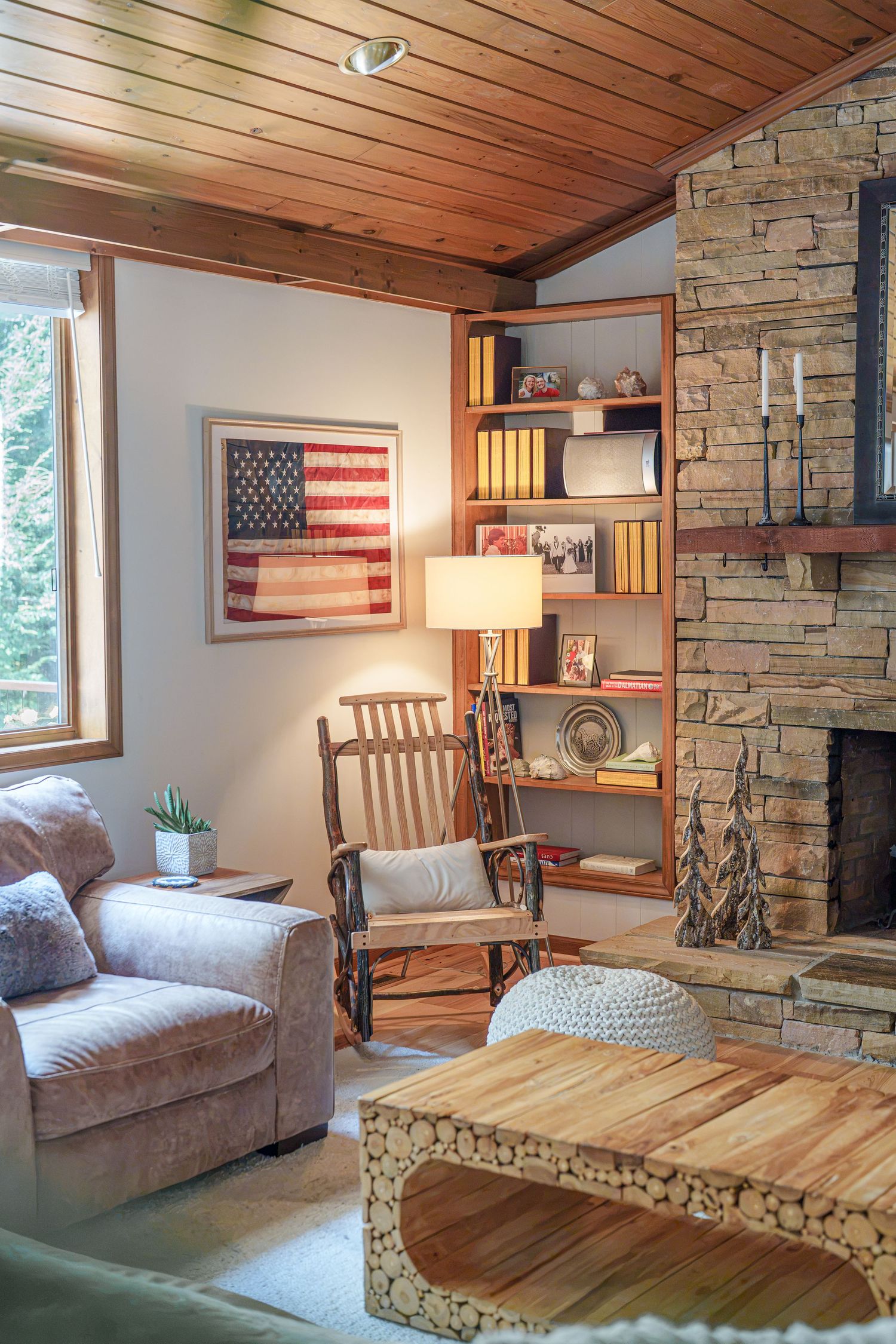 Cozy rustic living room with stone fireplace, wood ceiling, American flag art, rocking chair, and log coffee table.