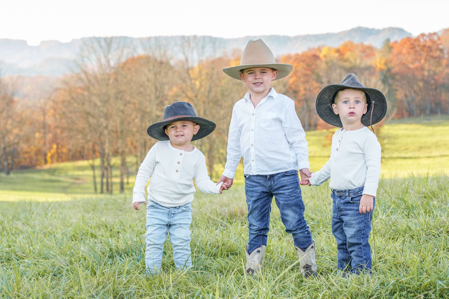 Three young boys in cowboy hats and white shirts hold hands in a green field at sunset.