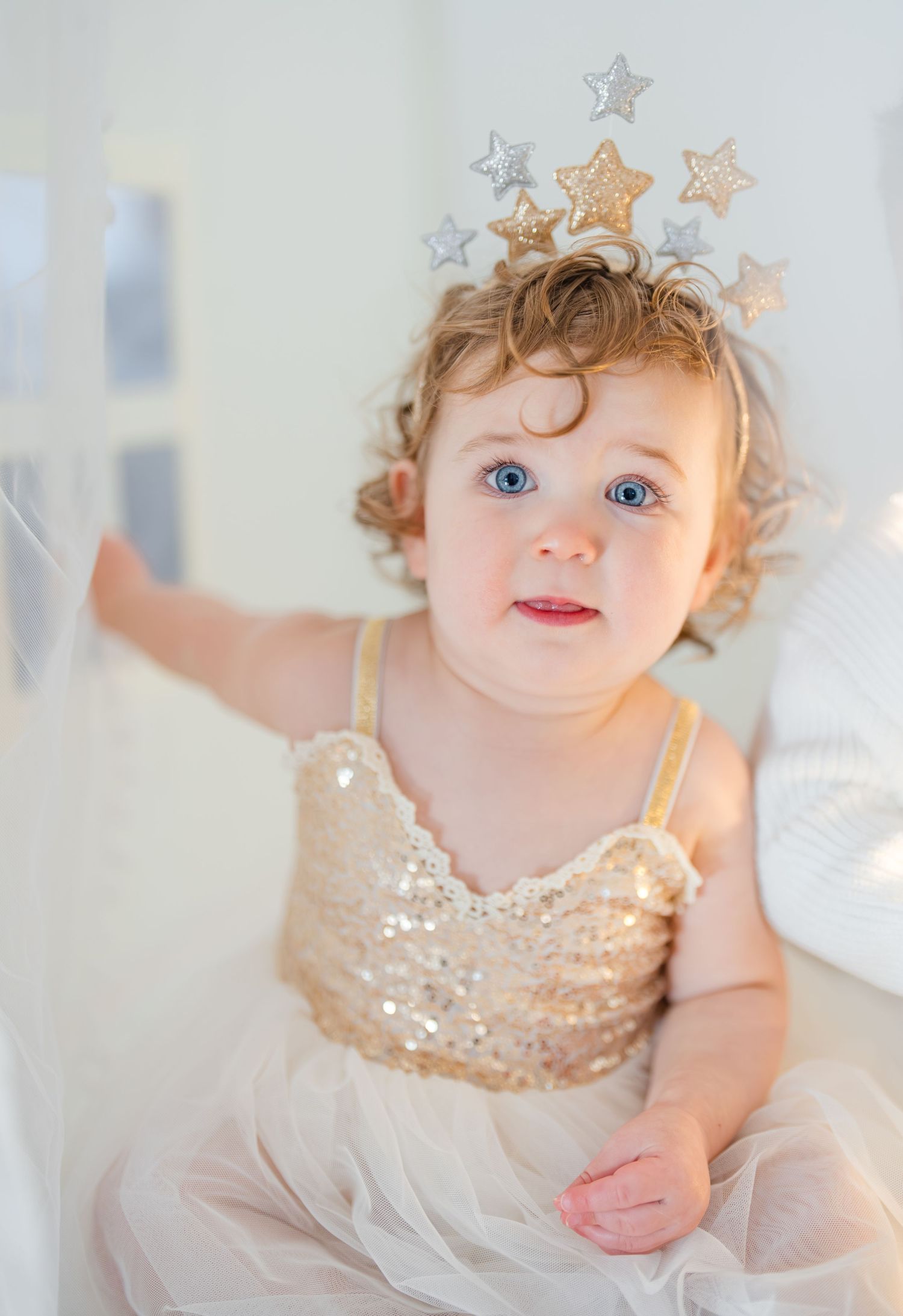 Adorable blue-eyed toddler girl in gold sequin dress wearing a star crown headpiece.