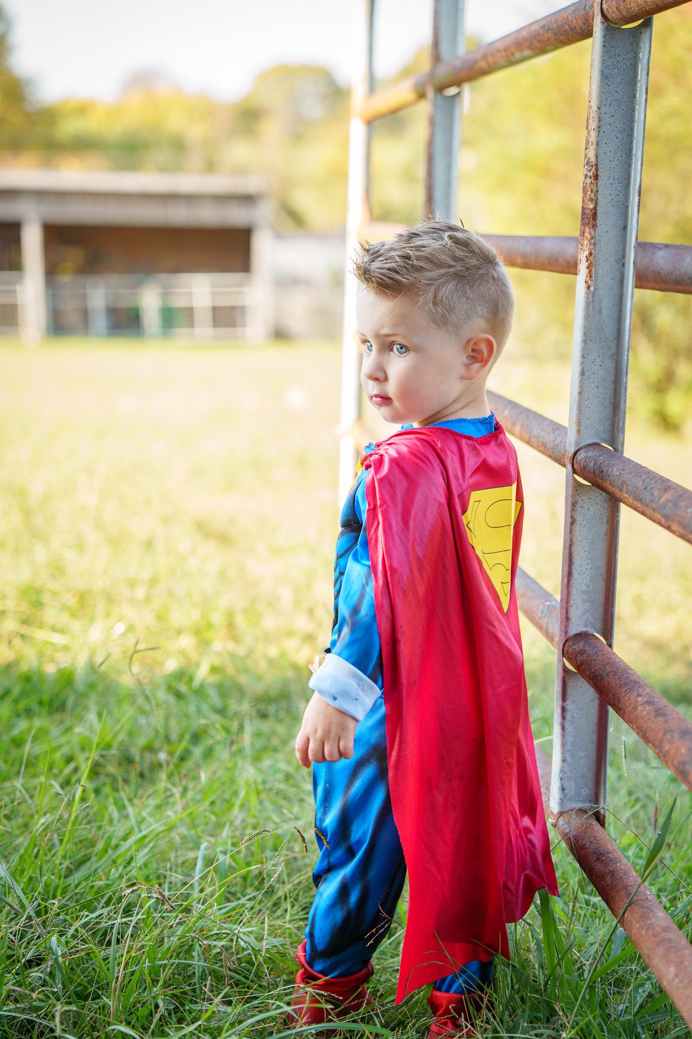 Young boy in Superman costume with red cape stands by rustic metal fence outdoors.