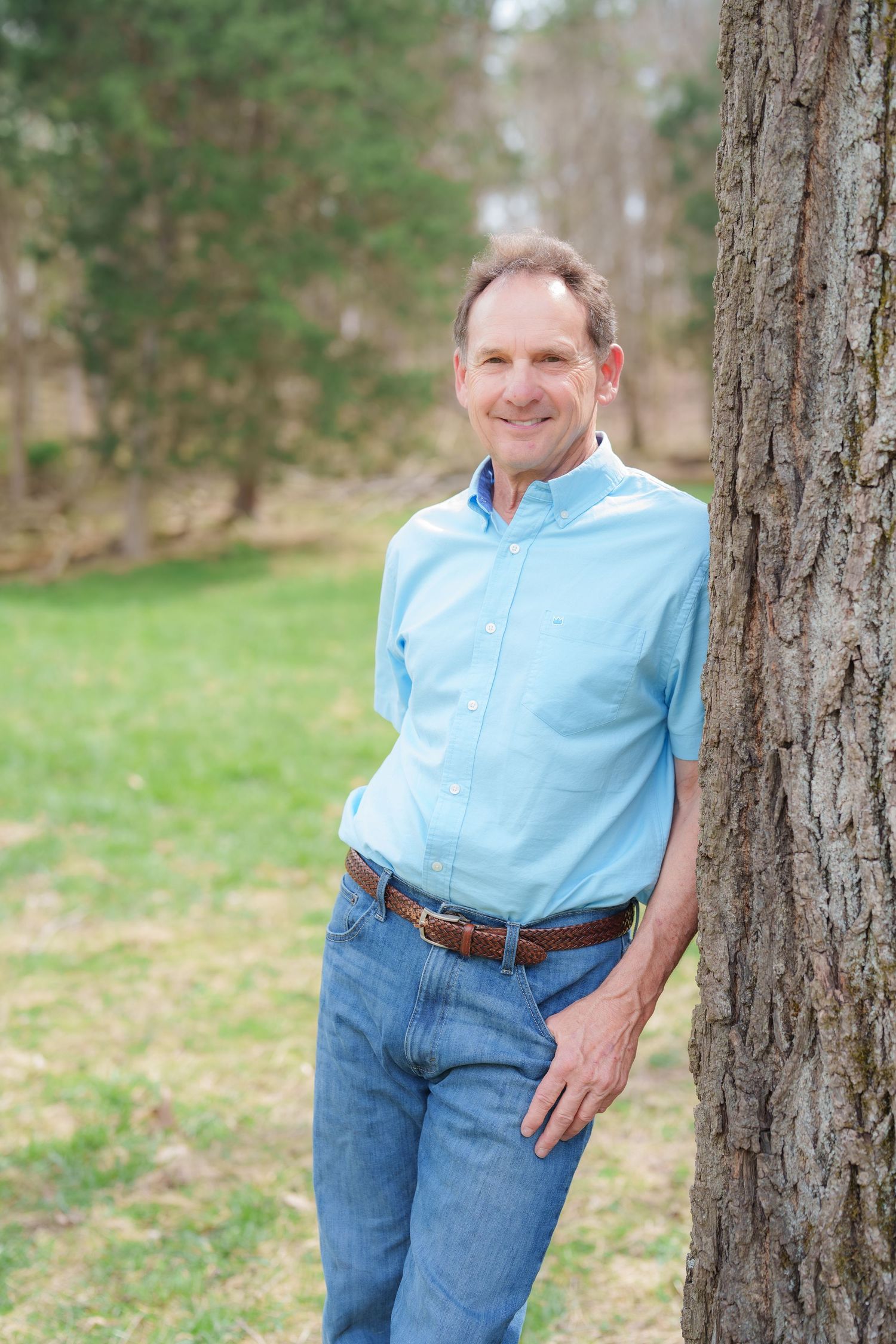 Middle-aged man in light blue shirt and jeans smiling while leaning against a tree in a park.
