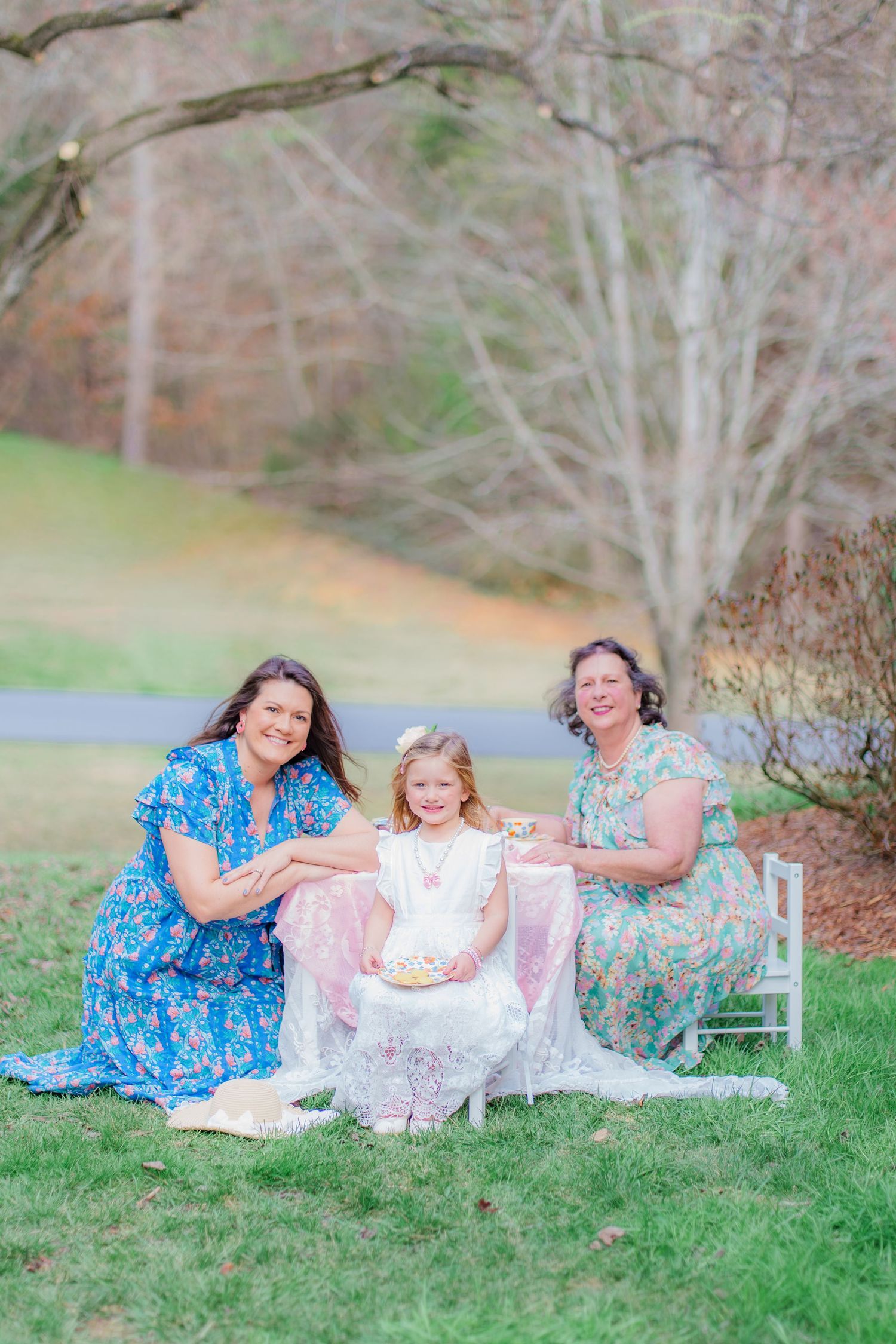 Three generations of women posing outdoors in floral dresses beside white chair in park.