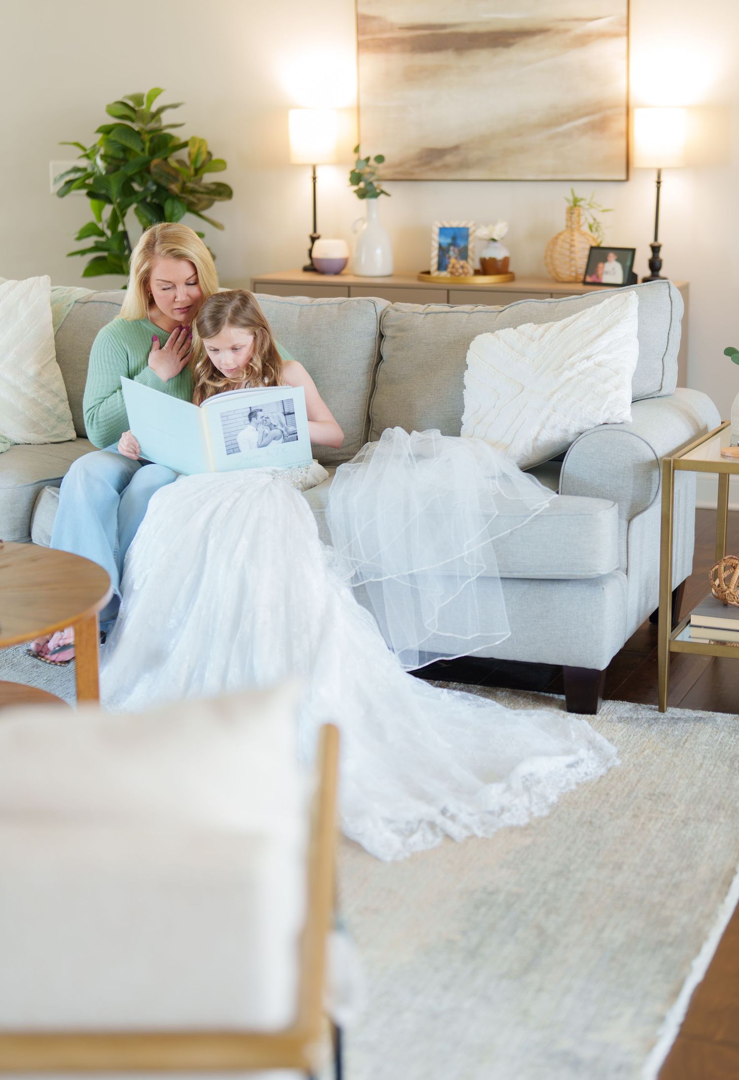 A blonde woman and young girl in a wedding dress sit on a gray sofa looking at a photo album together.