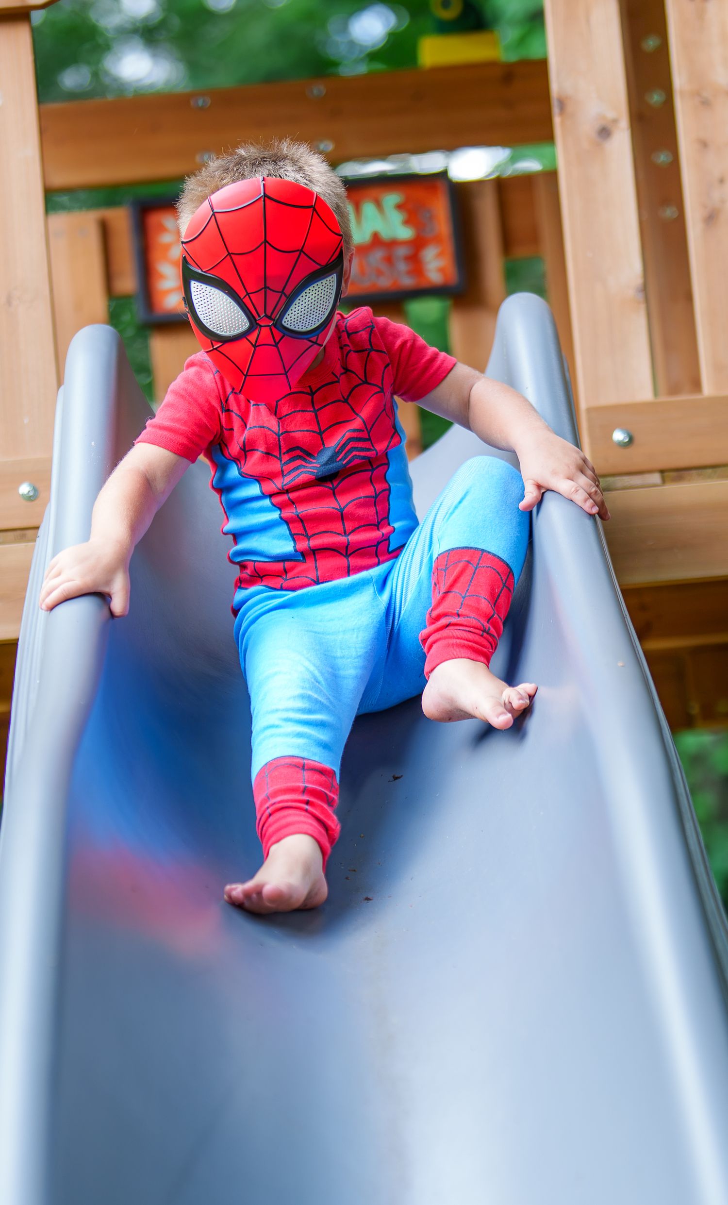 A young child wearing a Spider-Man costume slides down a playground slide barefoot on a sunny day.