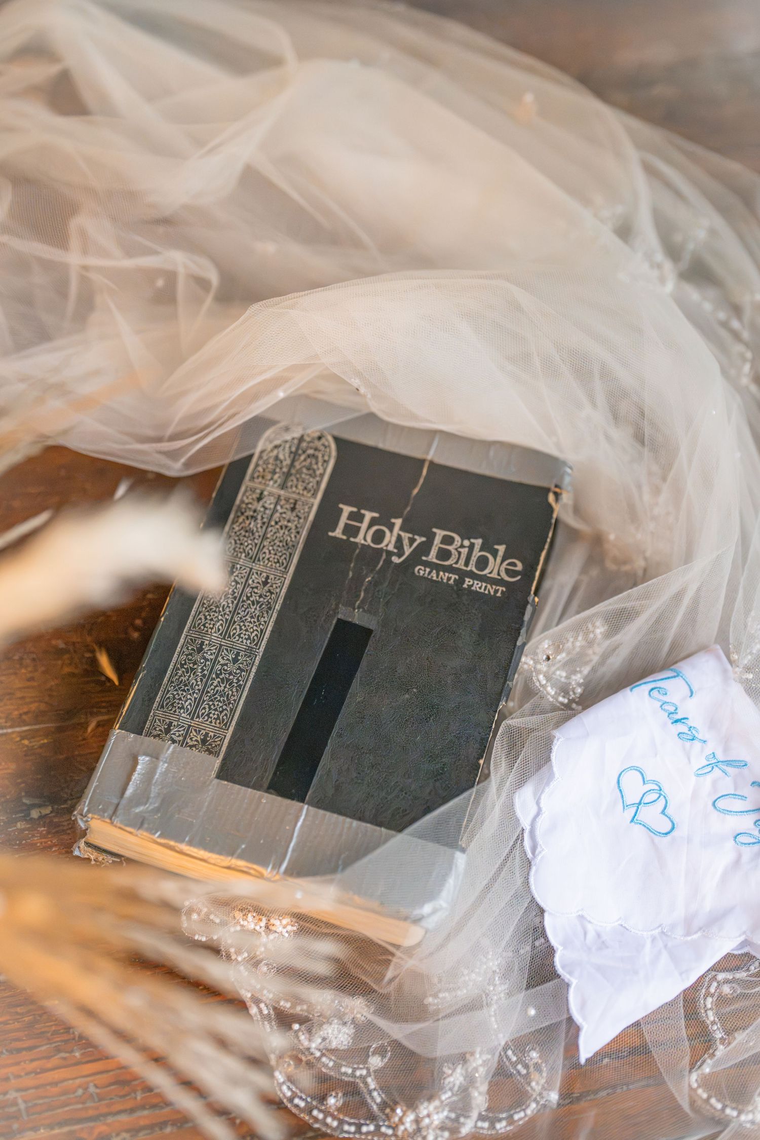 A worn Holy Bible with giant print edition rests beside a lace wedding veil and embroidered handkerchief on a wooden surface.