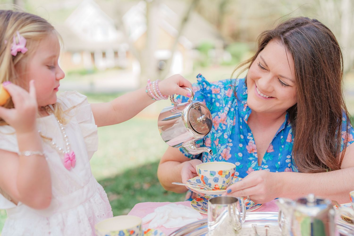 A woman smiles as a little girl pours tea at an outdoor tea party with floral teacups.