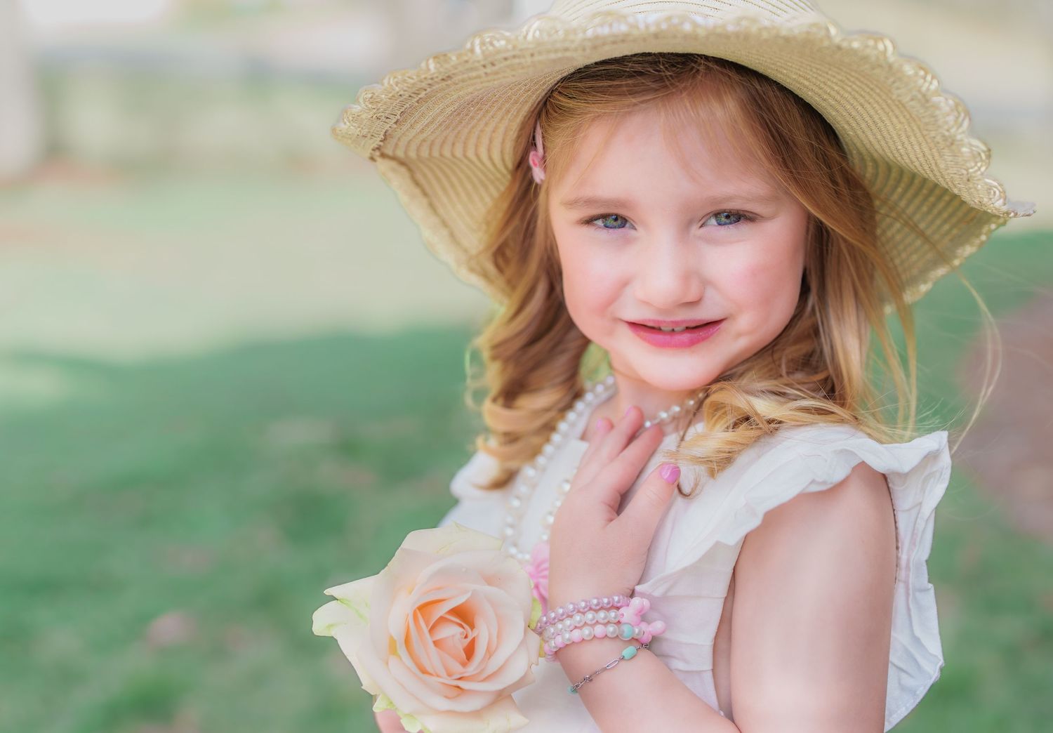 Young girl in straw hat holding peach rose, wearing white dress and pearl necklace, smiling outdoors.