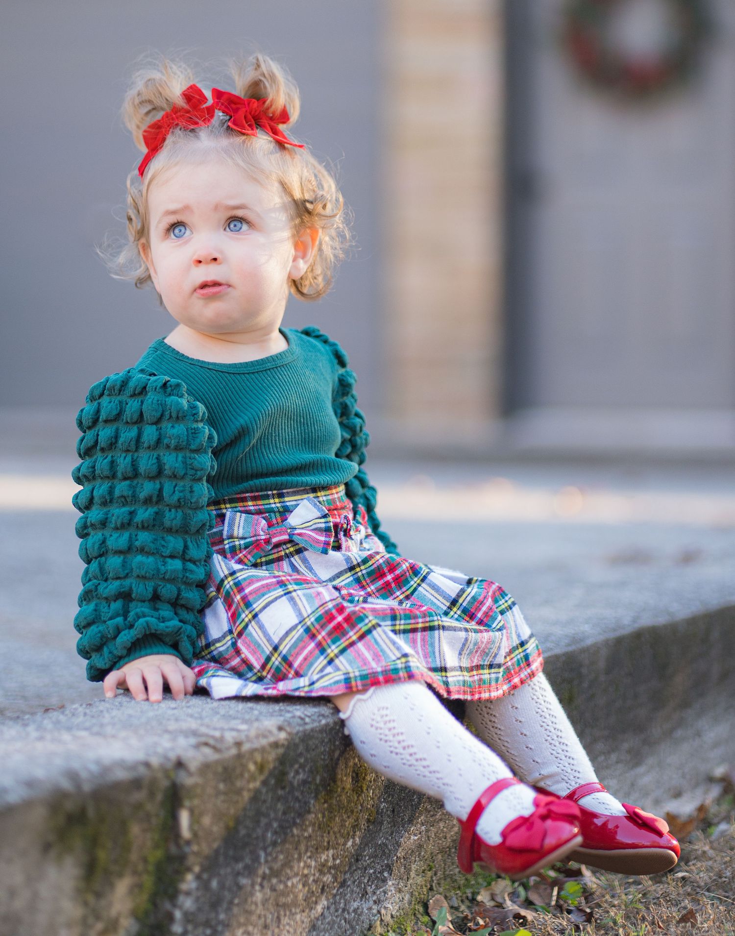 A toddler girl in a green bubble sweater and plaid skirt with red bow sits on stone steps outdoors.