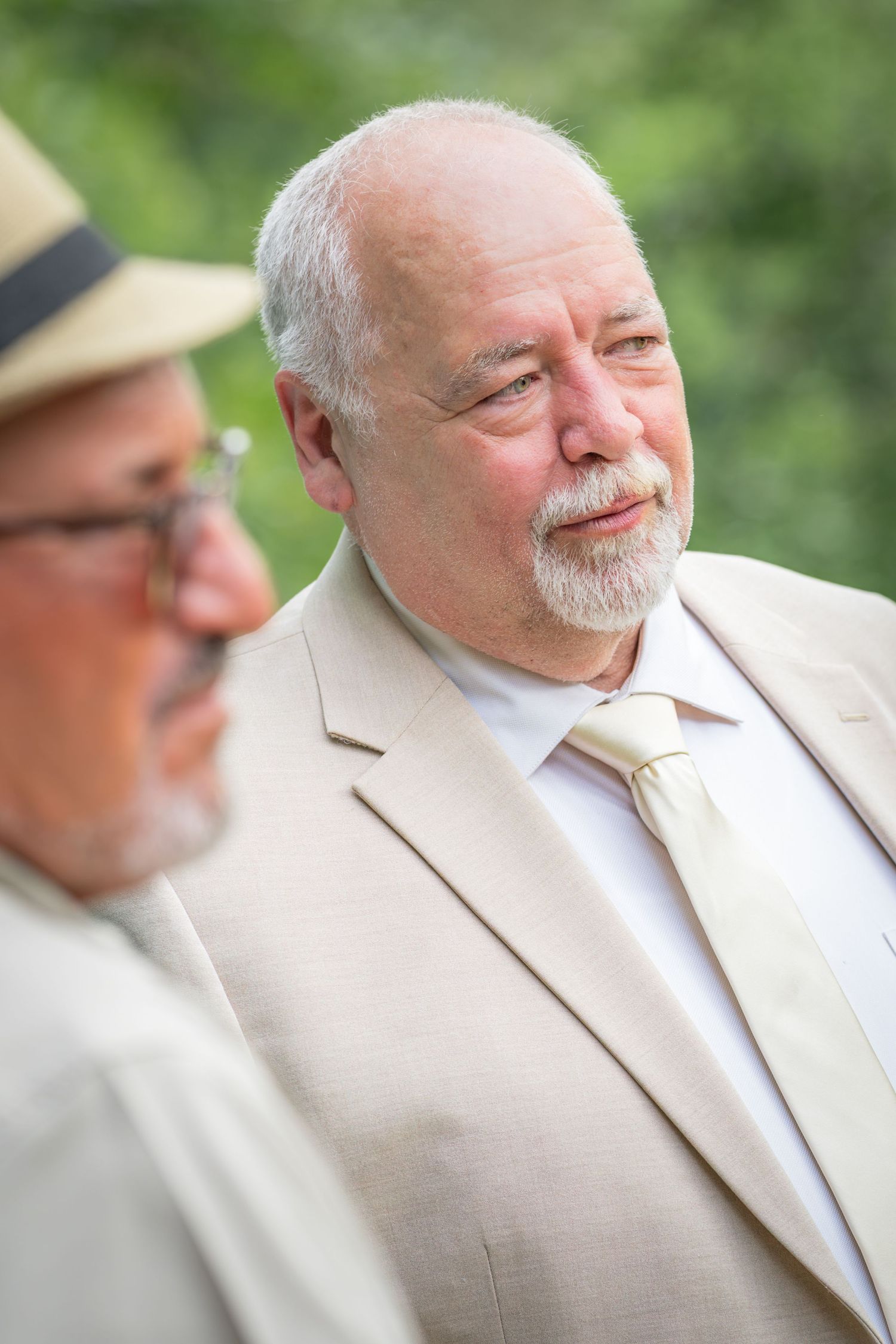 Two senior men in beige suits conversing outdoors, one bald with a goatee and tie, the other wearing a hat and glasses.