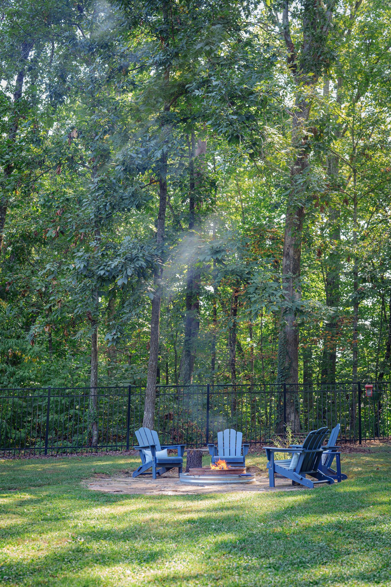 Backyard fire pit with blue Adirondack chairs surrounded by lush green trees and black metal fence.