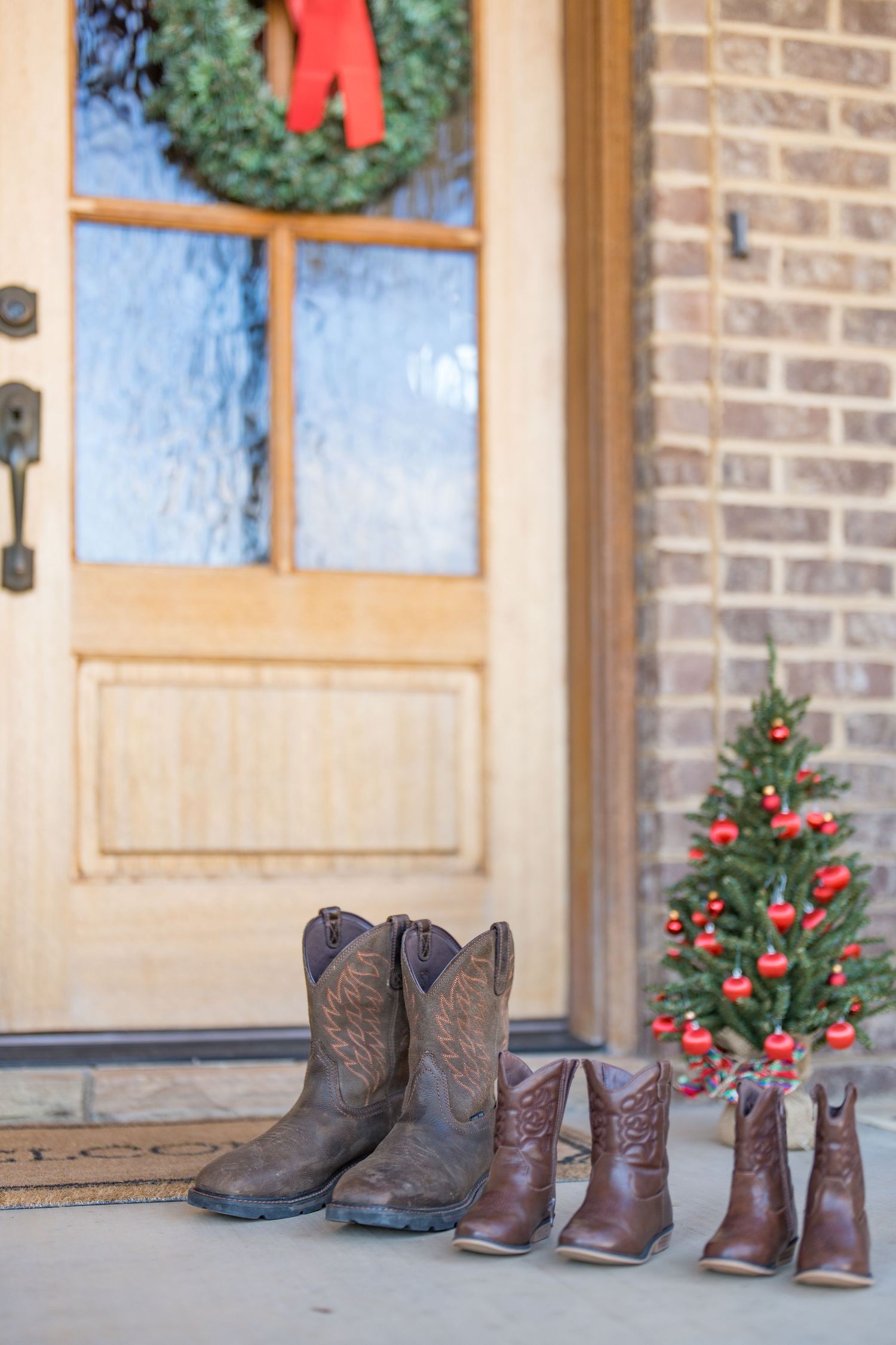 Three pairs of cowboy boots on a front porch with Christmas wreath and tree decorations.