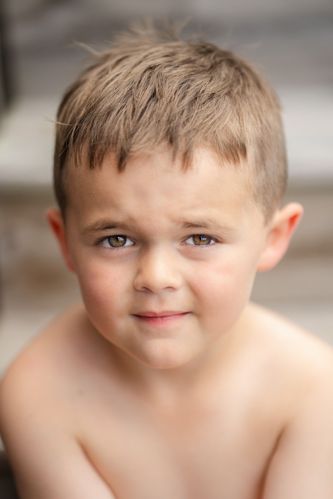 Close-up portrait of a young shirtless boy with hazel eyes and a subtle smile outdoors.