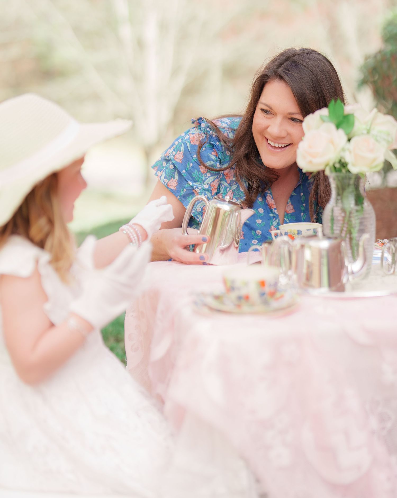 A smiling woman in a blue floral dress shares a tea party with a young girl outdoors.