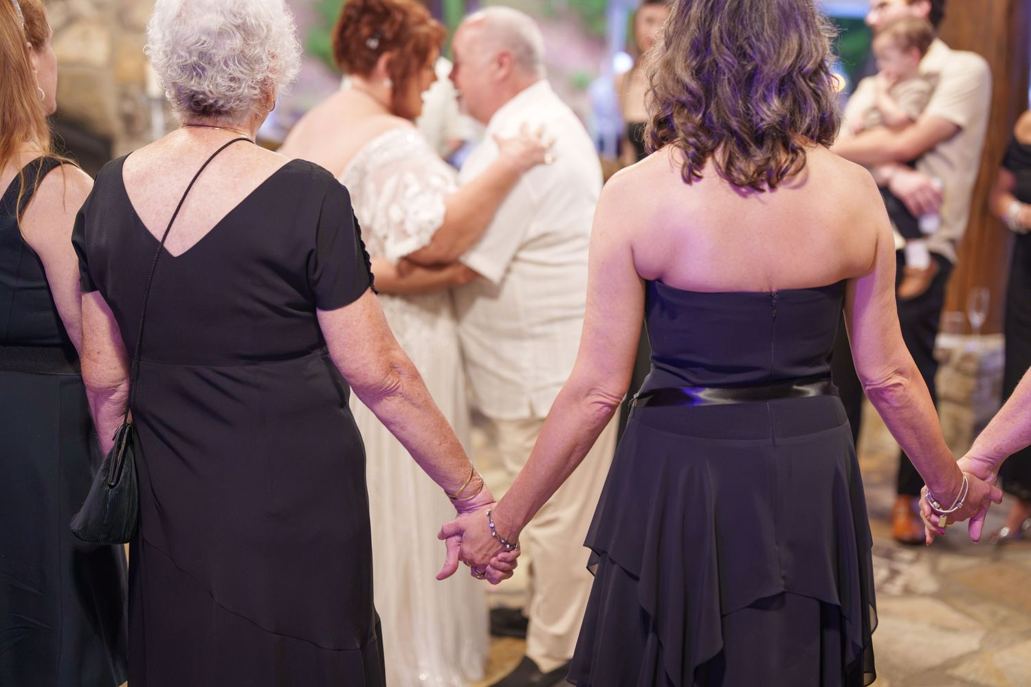 Two women in black dresses hold hands at a wedding reception while guests dance in the background.