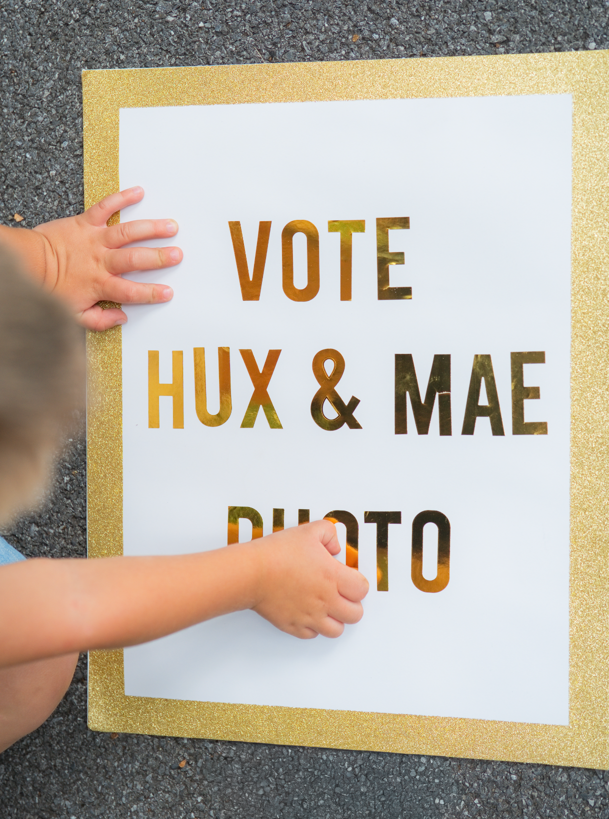 Child holding a white poster with gold glitter border reading Vote Hux & Mae Duo, perfect for a campaign or election event.