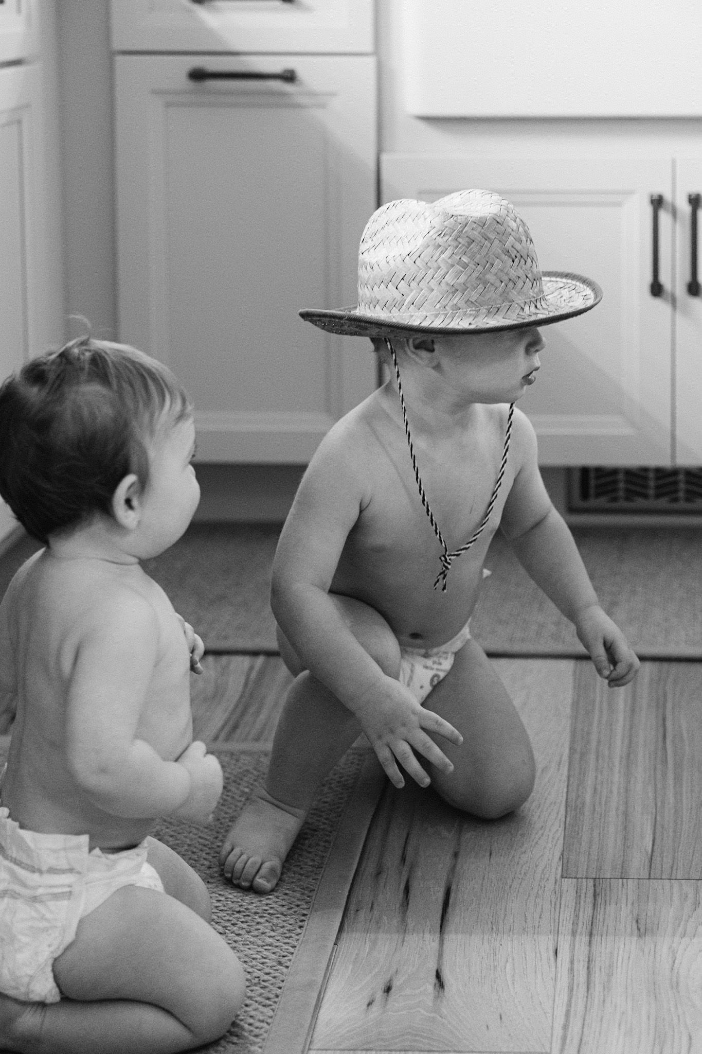 Black and white photo of two babies in diapers sitting on a kitchen floor, one wearing a straw hat.