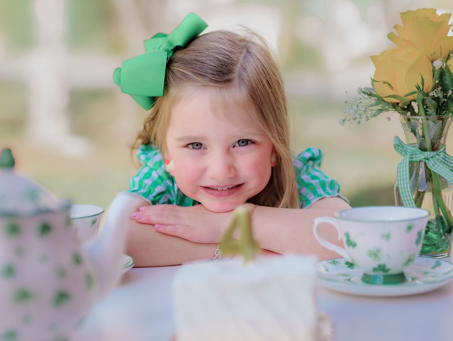 A sweet young girl in a green gingham dress smiles while leaning on a table set with tea and cake.
