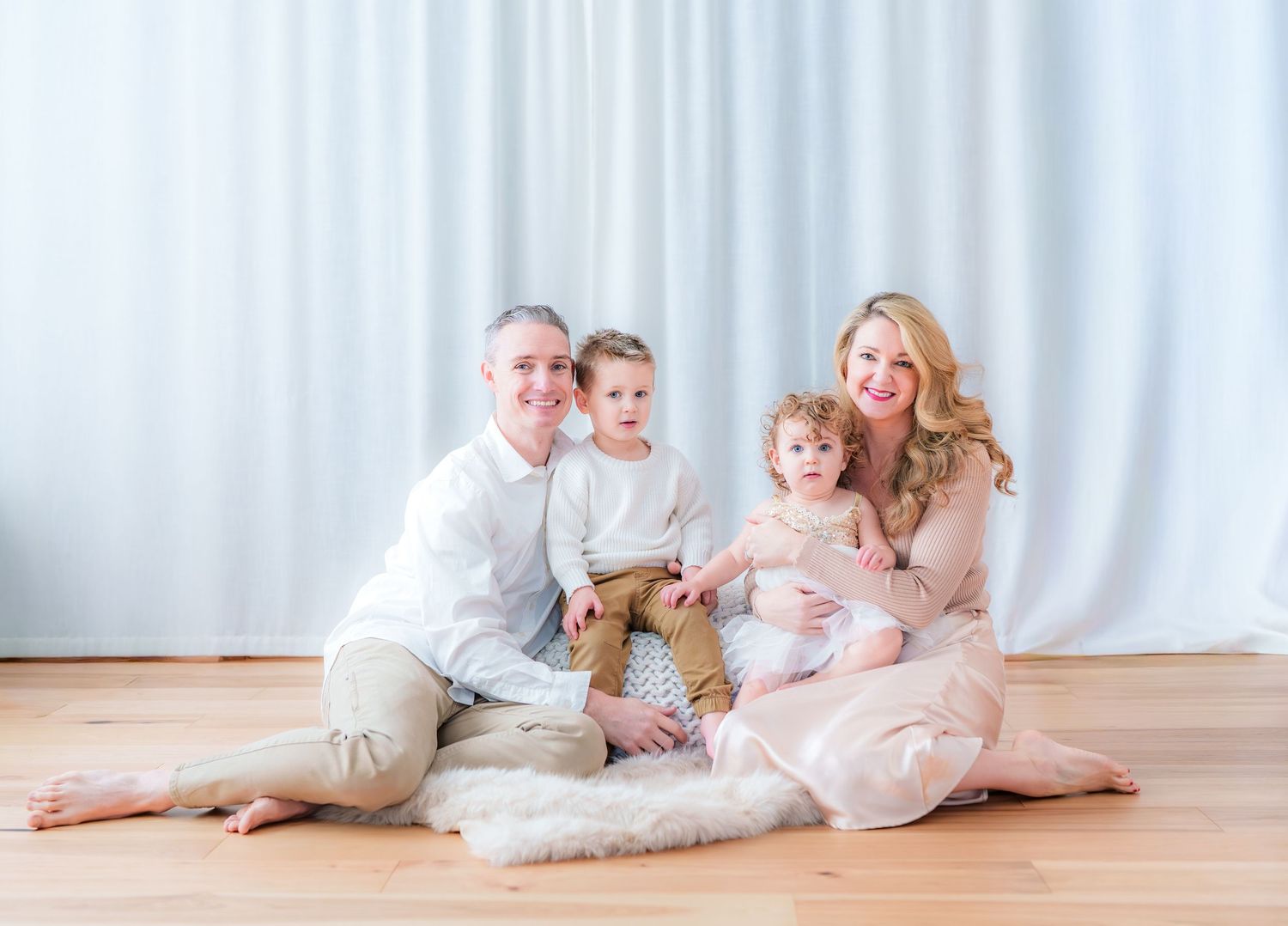 Happy family of four sitting on a fur rug against white curtains during a professional photo session.