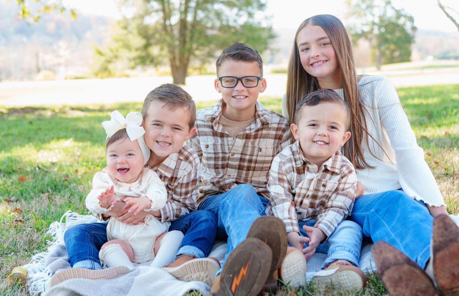 Five siblings in matching plaid shirts smile together while sitting on a blanket in a sunny outdoor setting.