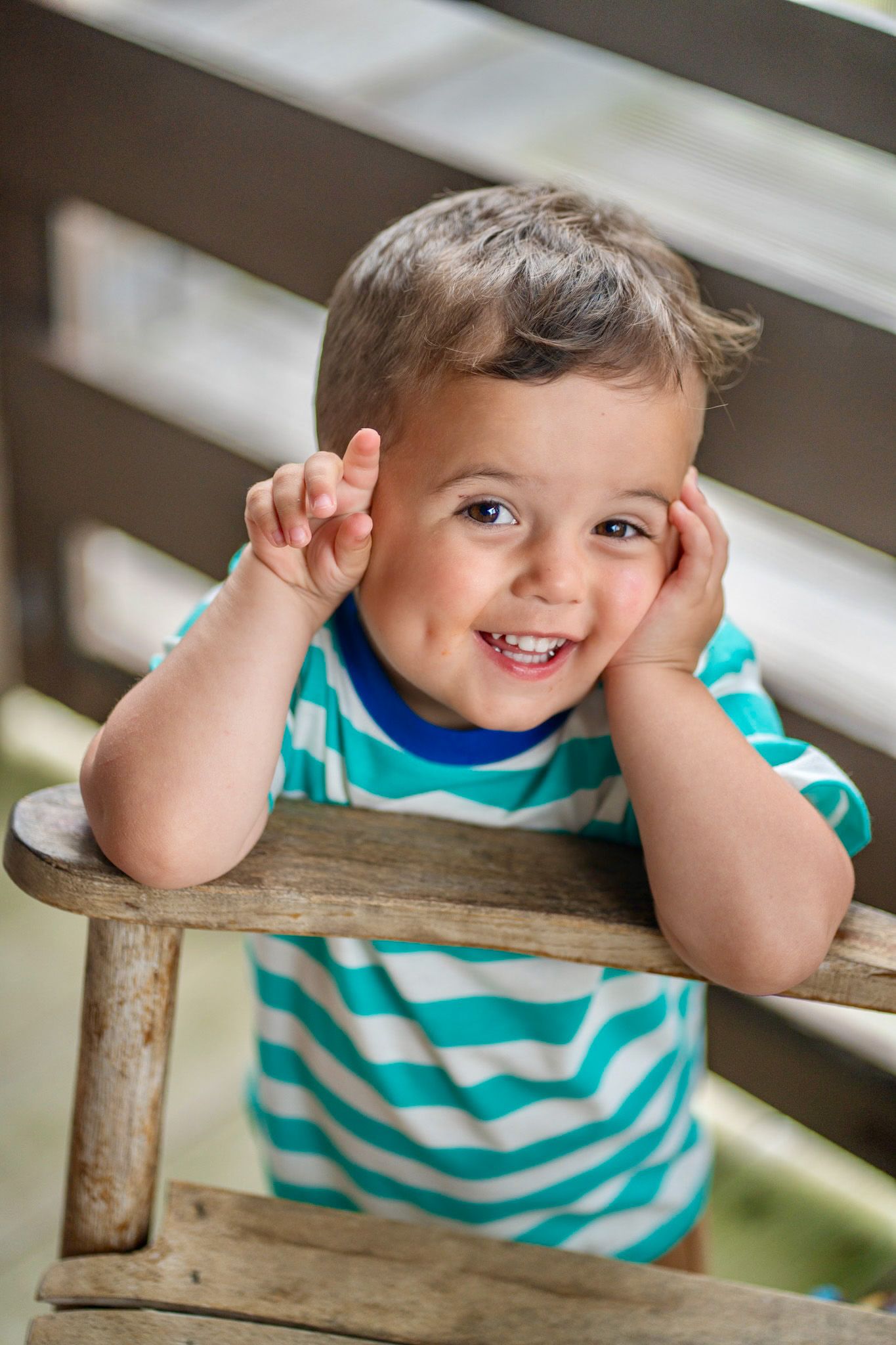 Adorable smiling toddler boy in striped shirt leans on wooden chair railing with hands on cheeks.