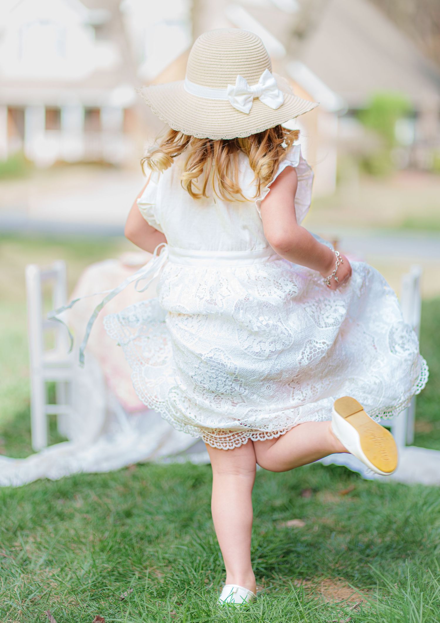 A little girl in a white lace dress and sun hat twirls joyfully on green grass.