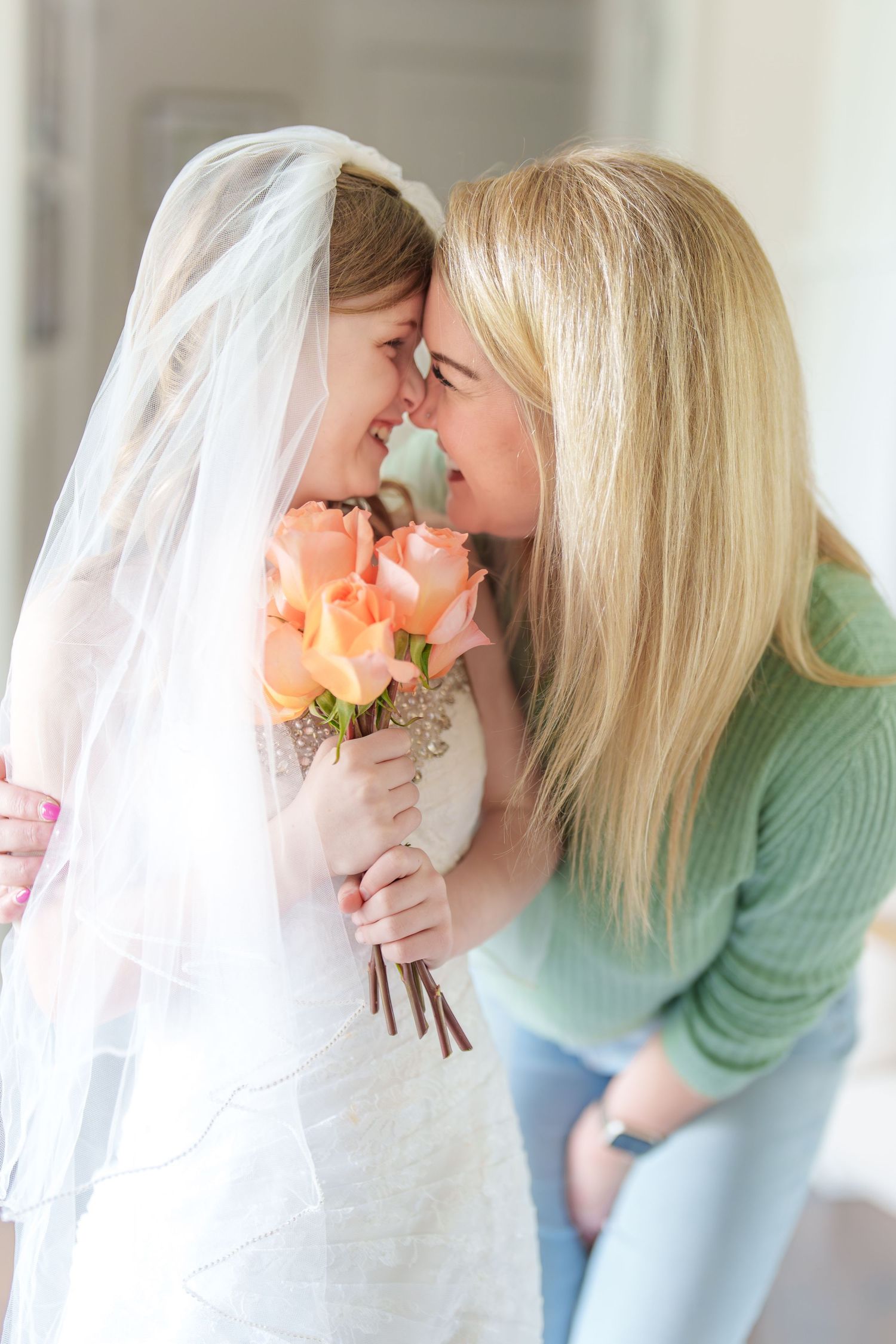 A bride in white veil and blonde woman touch foreheads while holding peach roses bouquet.