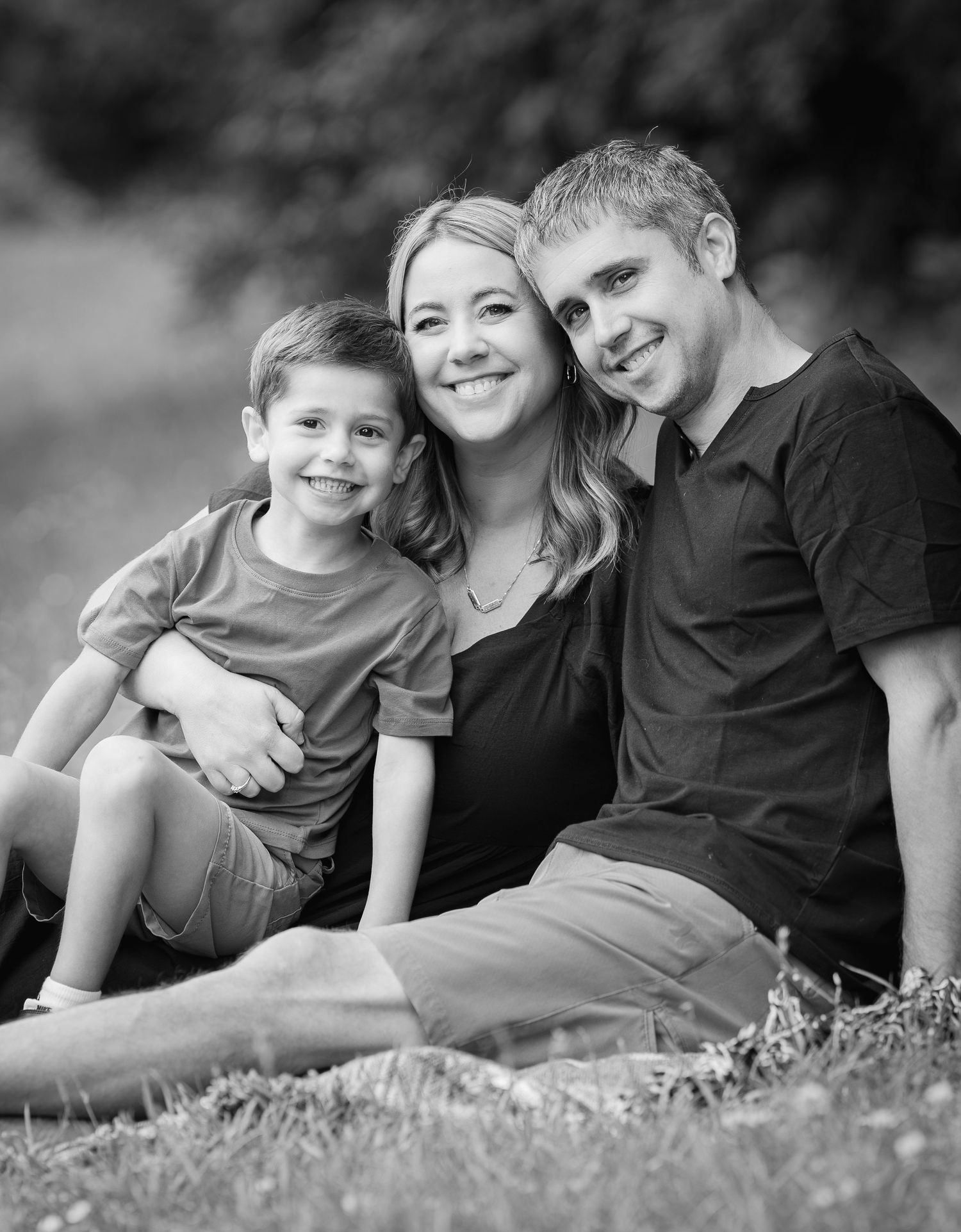 Black and white family portrait of smiling mother, father, and young son sitting together on grass outdoors.