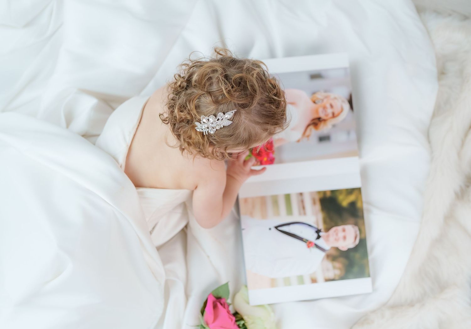Curly-haired toddler girl with tiara looking at wedding photo albums on a white bed.