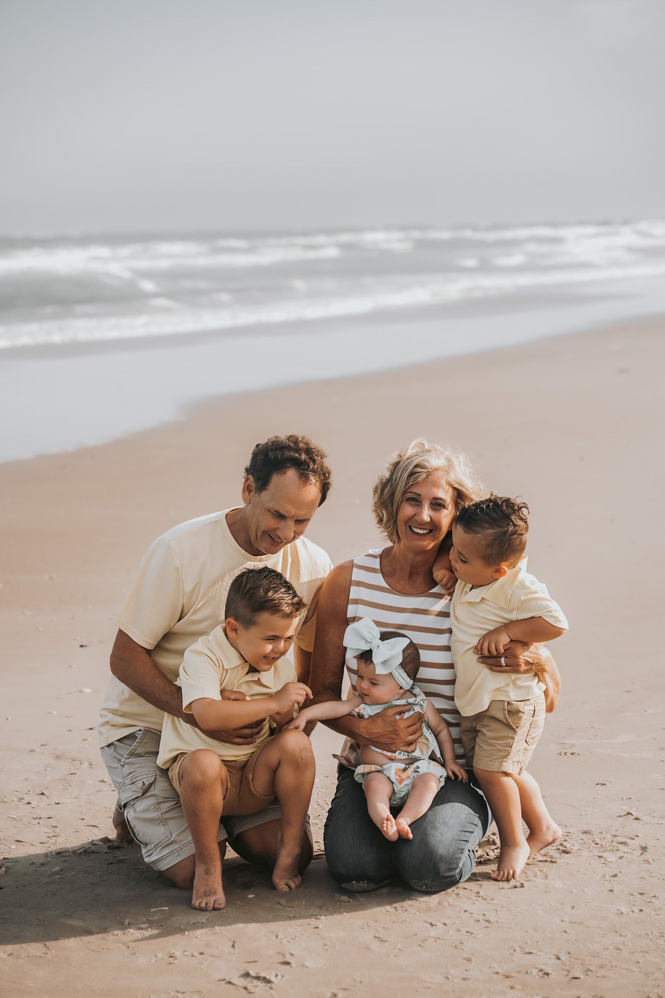 Grandparents sit on a beach with three young grandchildren, smiling together near ocean waves.