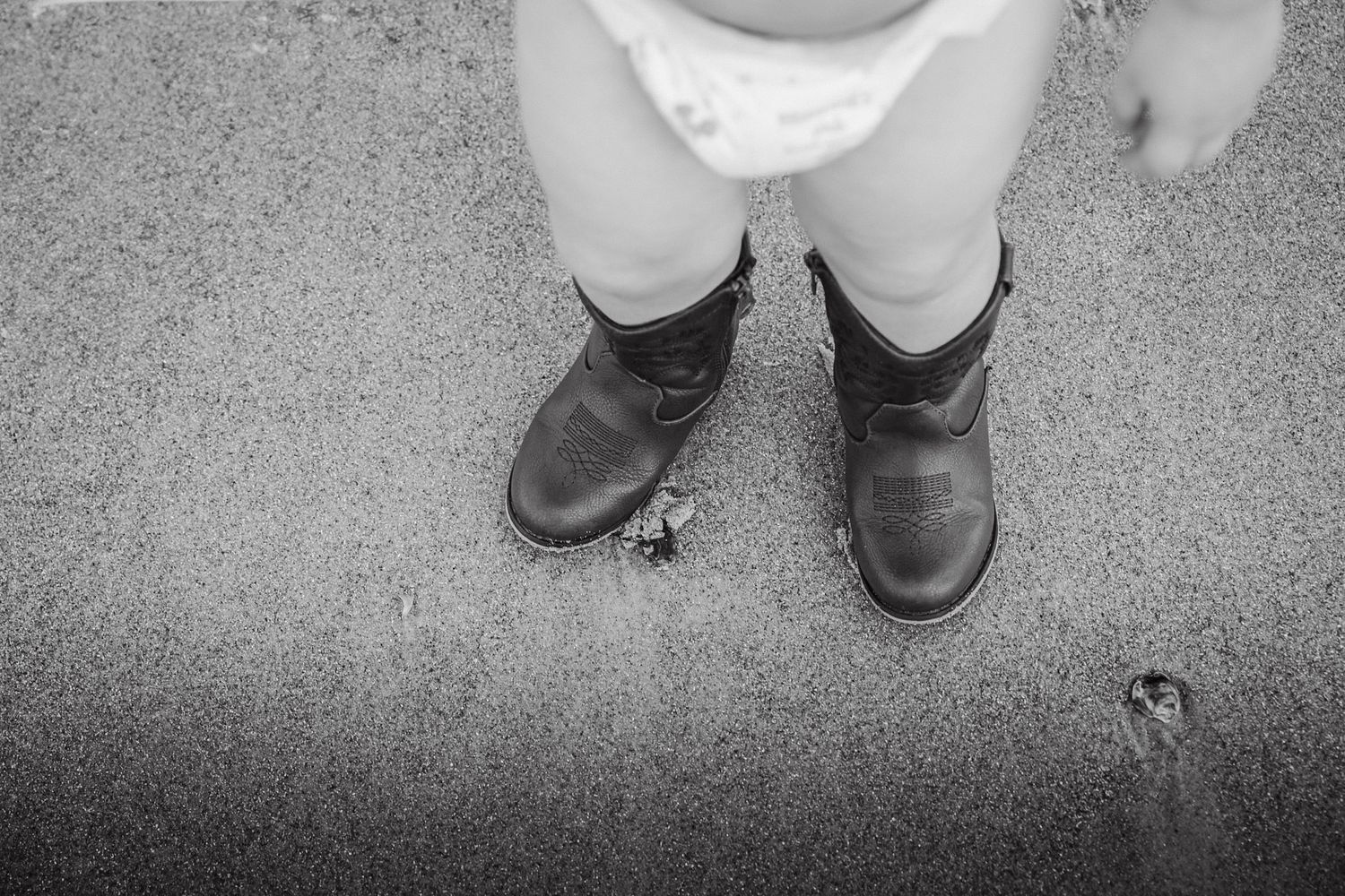 Close-up of toddler feet wearing brown cowboy boots on sandy beach.